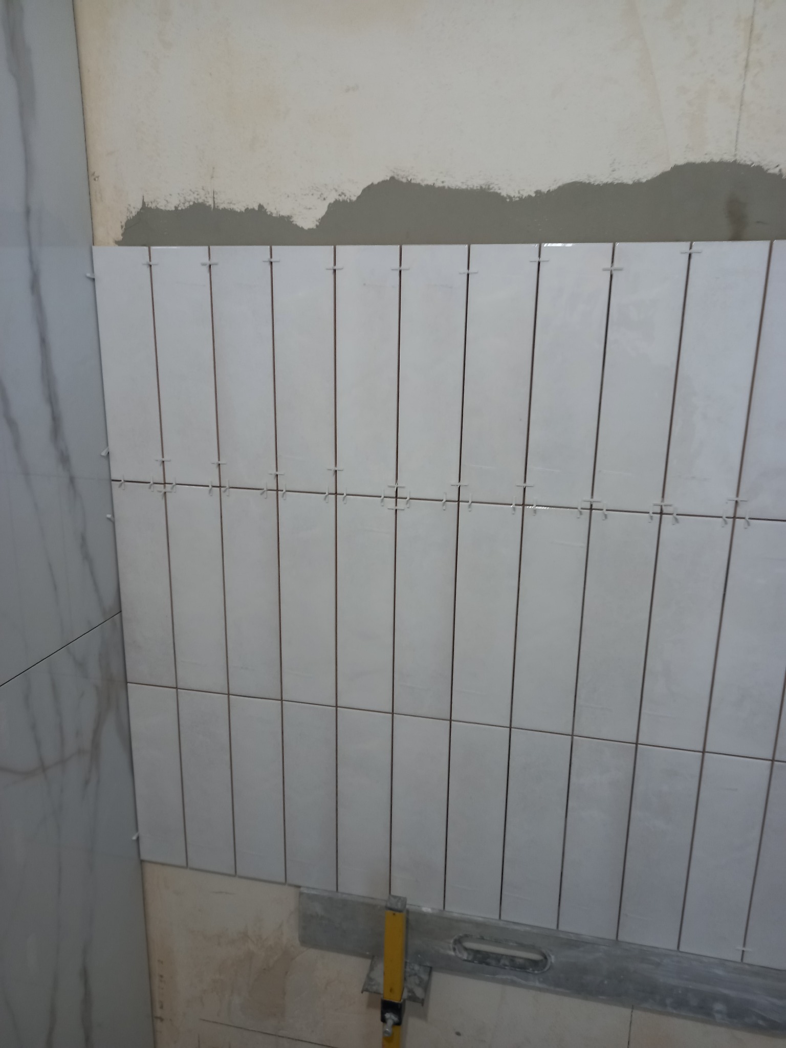 Układanie wąskich, białych płytek na ścianie z użyciem krzyżyków dystansowych. Widoczny fragment ściany przygotowanej pod glazurę i poziomica.