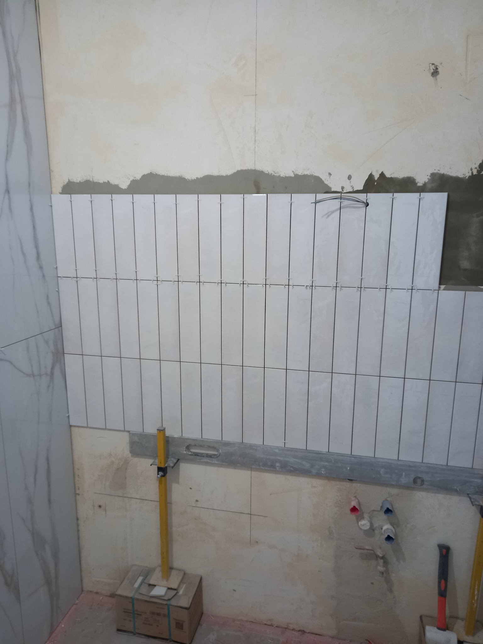 Układanie białych, podłużnych płytek na ścianie w łazience, widoczne poziomice i krzyżyki dystansowe, przygotowane podłoże.