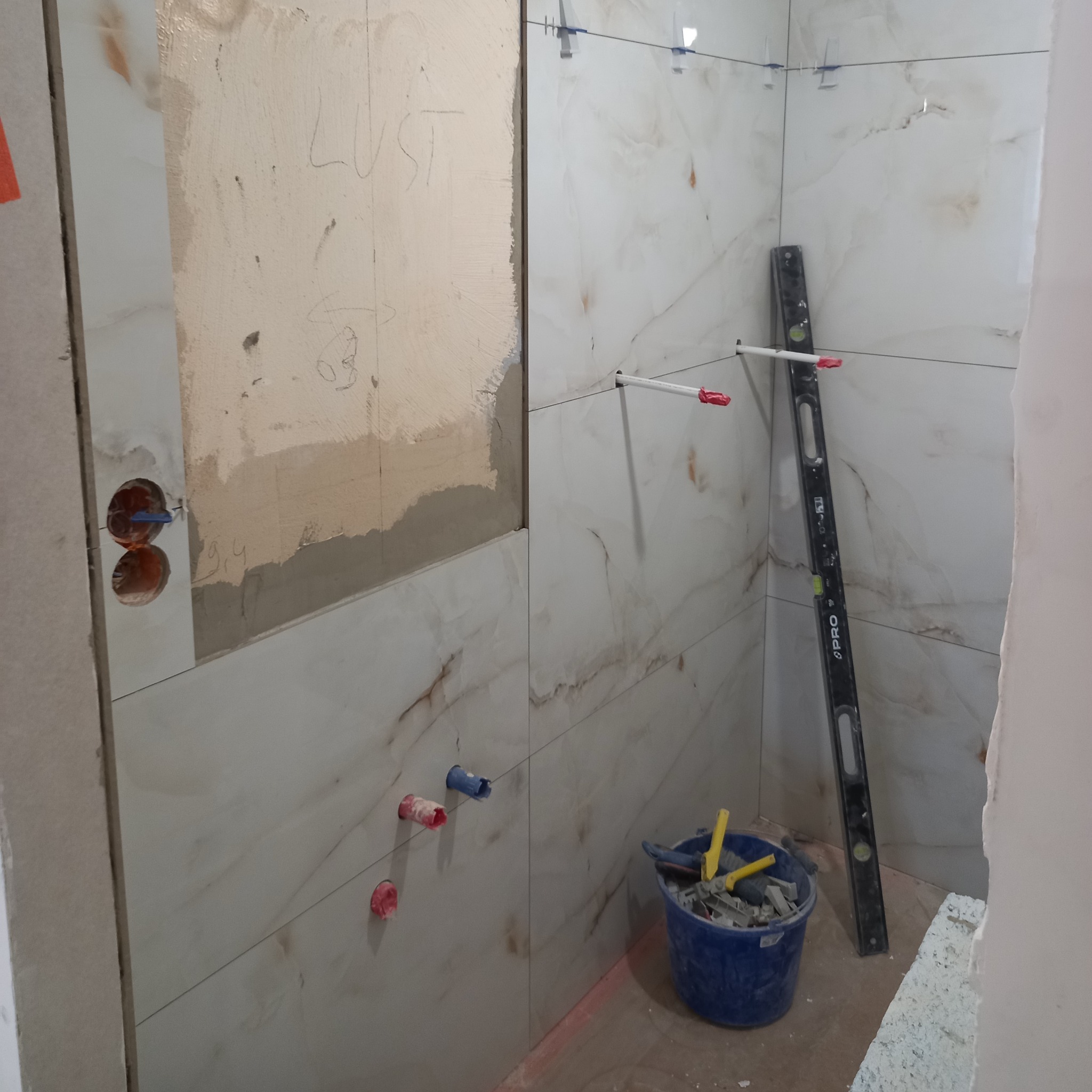 Montaż płytek imitujących marmur w łazience. Widoczny poziomica, wiadro z narzędziami i fragment ściany z napisem 'LUST'.