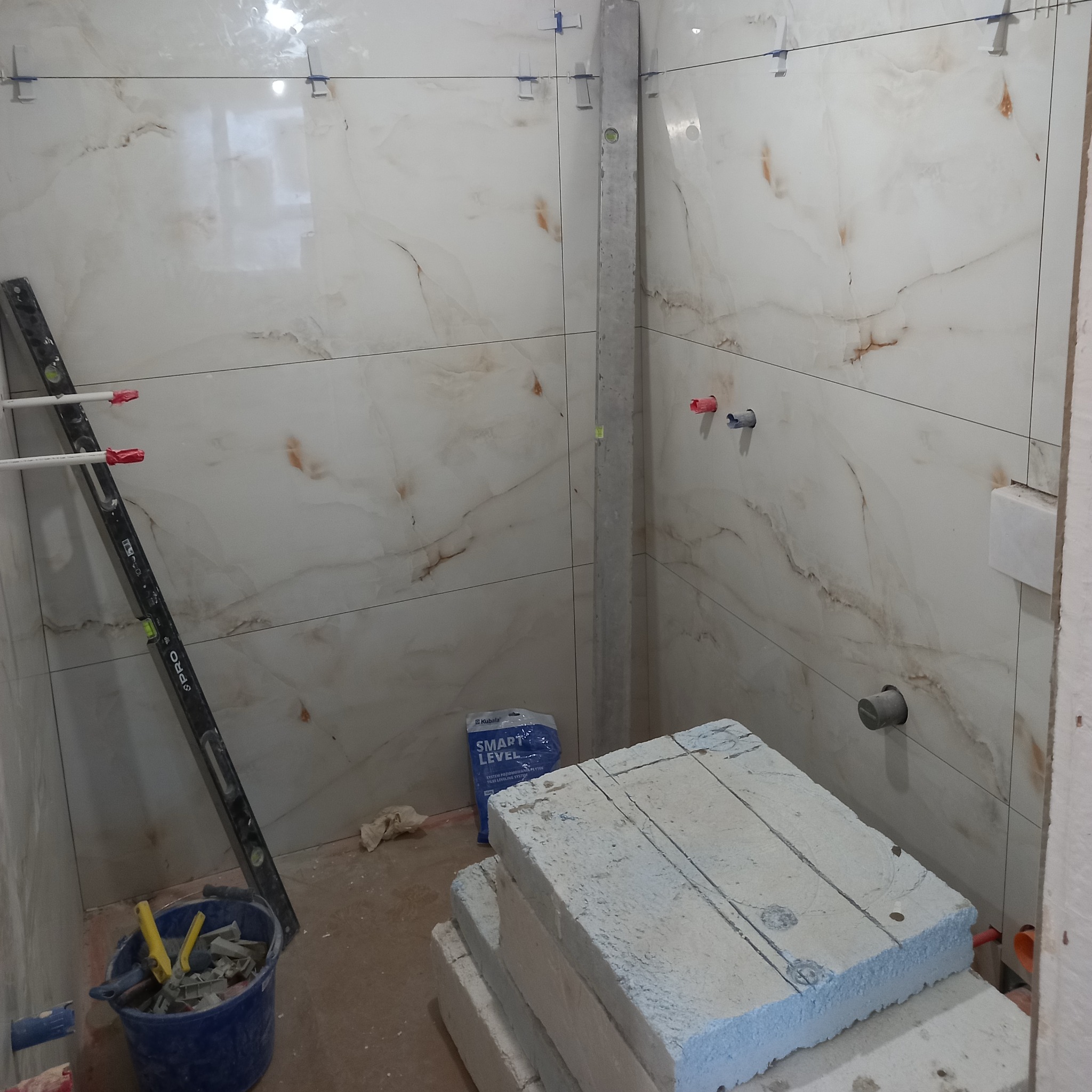 Ściana w trakcie układania płytek imitujących marmur, poziomica i narzędzia glazurnicze w wiadrze, worek z systemem poziomowania płytek.