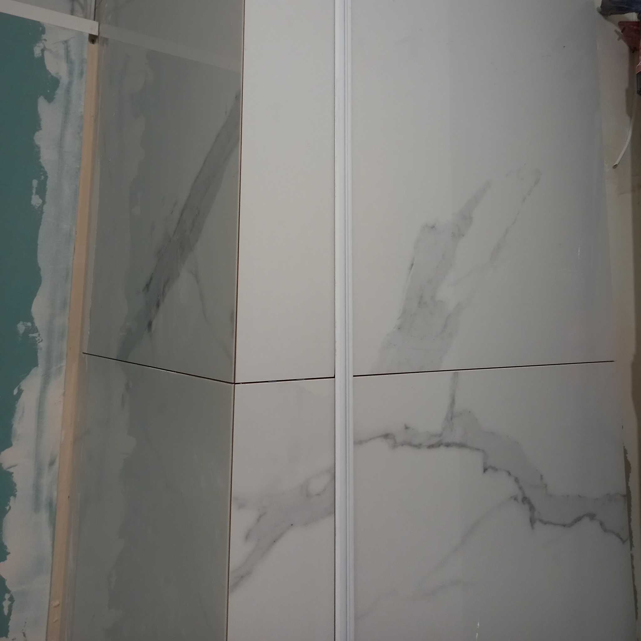 Fragment ściany z białych płytek imitujących marmur, ułożonych w regularny wzór. Widoczne fugi i narożnik z profilem wykończeniowym. Częściowo widoczna surowa ściana.