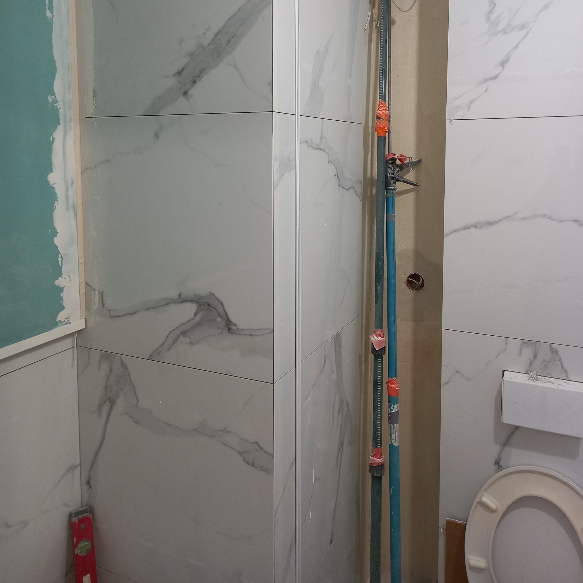 Białe płytki imitujące marmur w trakcie montażu w łazience. Widoczny poziomica, podpórka teleskopowa i fragment toalety. Ściana częściowo przygotowana pod płytki.