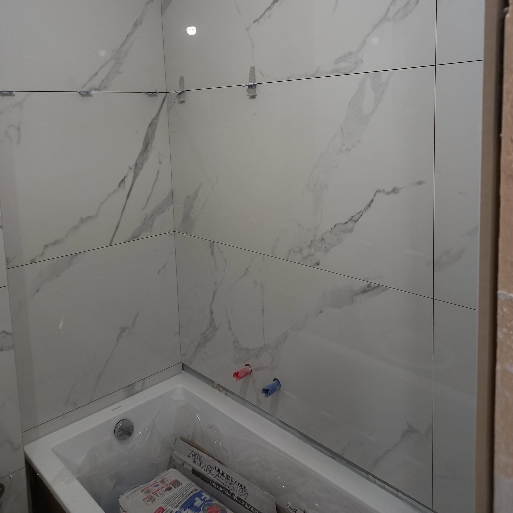Ściana łazienki z białymi płytkami imitującymi marmur, z widocznymi krzyżykami glazurniczymi, nad wanną. Częściowo widoczne opakowania płytek.