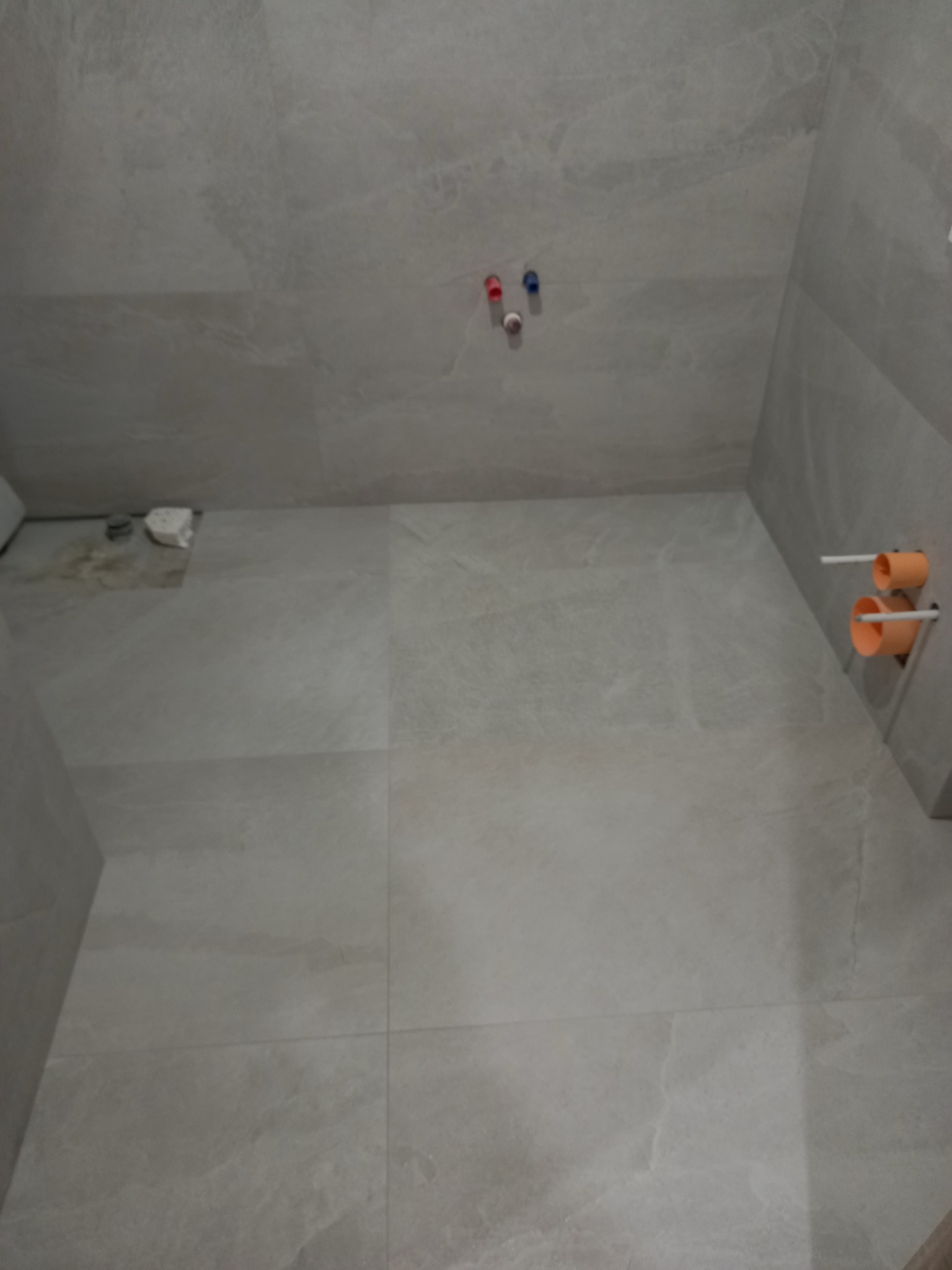 Wykończona, jasnoszara łazienka w trakcie prac: ułożone płytki podłogowe i ścienne, widoczne przyłącza wodne i kanalizacyjne. Minimalistyczny design.