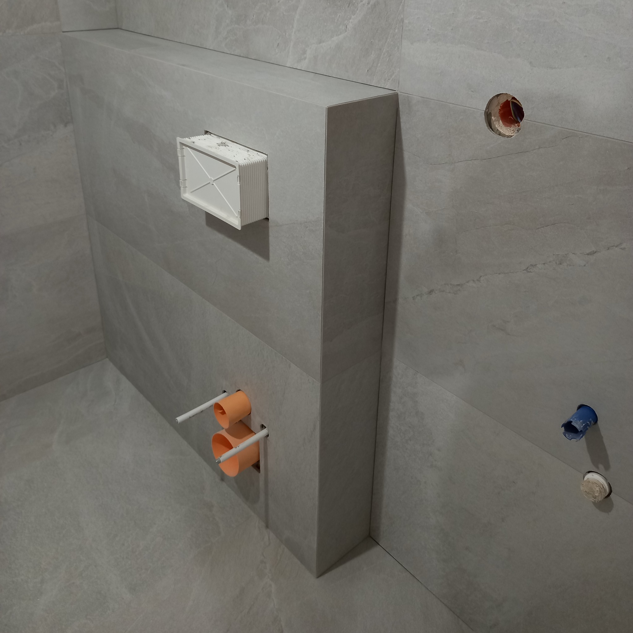 Szara, wykafelkowana łazienka w trakcie wykańczania. Widoczne otwory na rury i instalacje, oraz stelaż podtynkowy WC. Minimalistyczny styl.