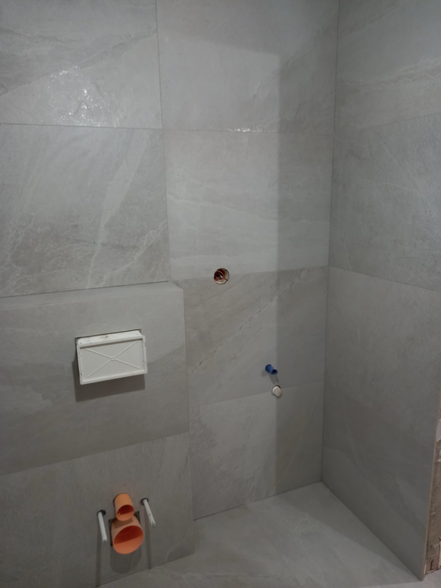 Wykończona łazienka z szarymi płytkami imitującymi marmur. Widoczne elementy instalacji wod-kan i stelaż podtynkowy. Minimalistyczny design, surowy styl.