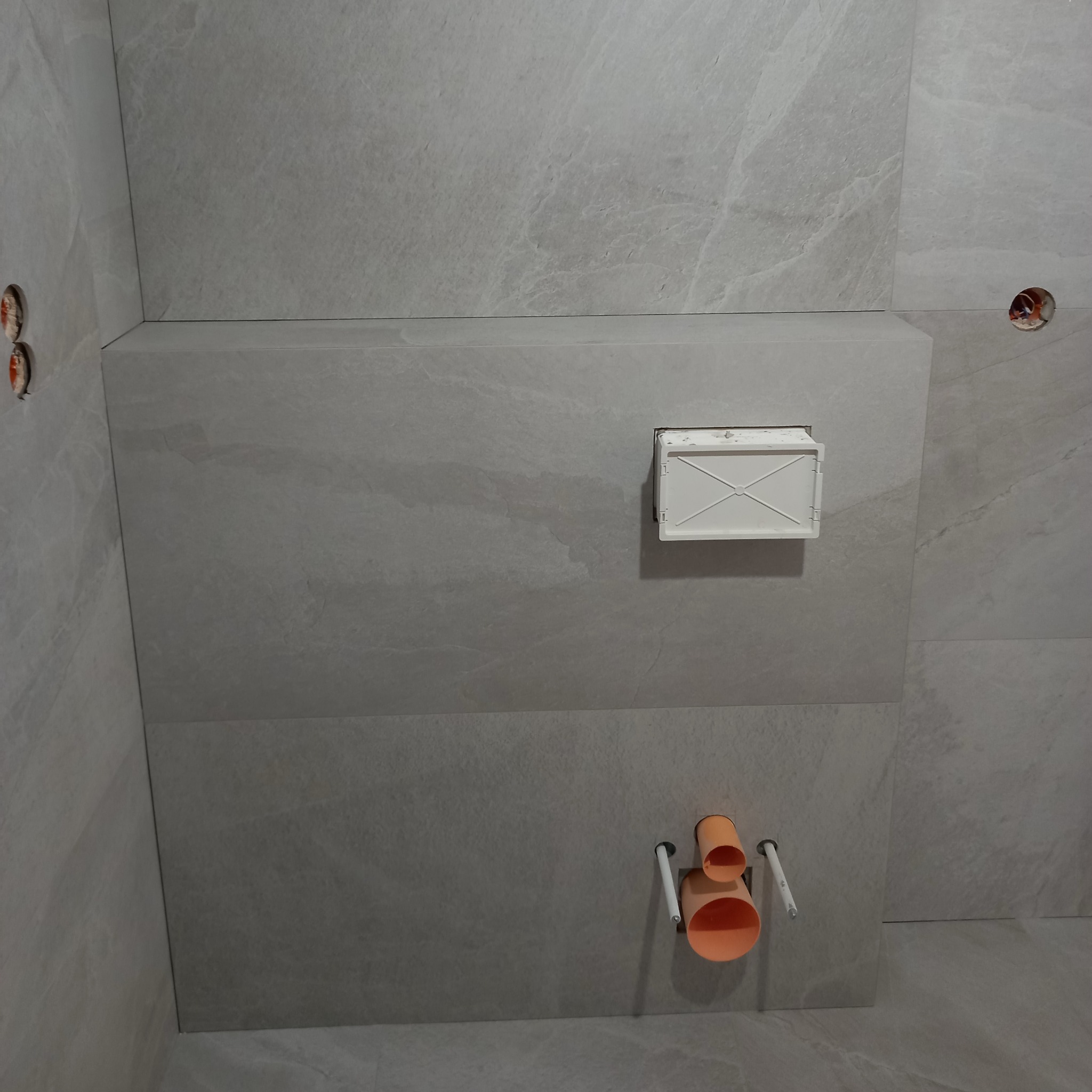 Wykończona ściana łazienki z szarymi płytkami imitującymi kamień, widoczne rury i stelaż podtynkowy WC. Minimalistyczny design.