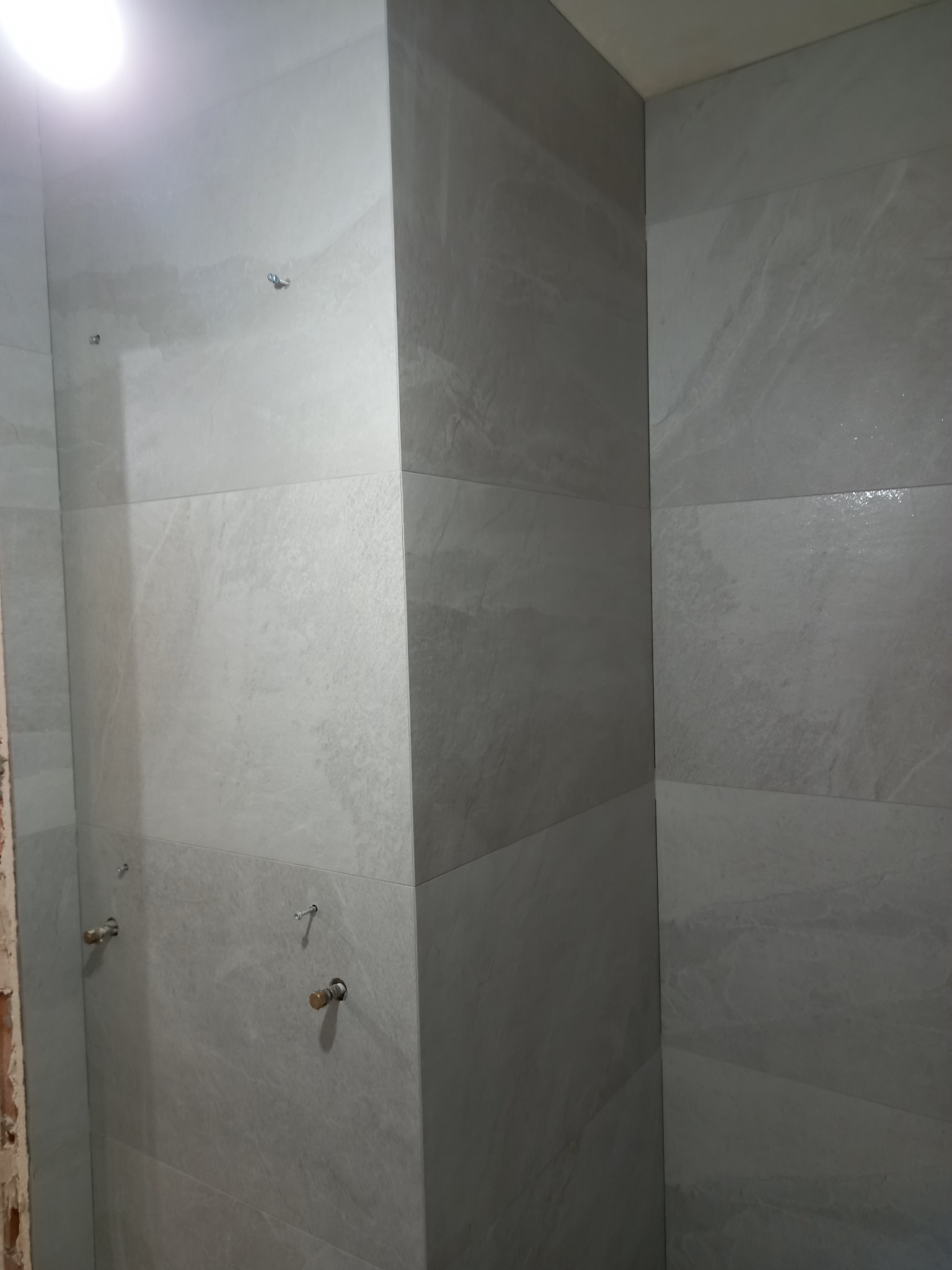 Wykończona ściana w łazience z szarych, matowych płytek imitujących kamień. Widoczne mocowania baterii. Minimalistyczny design z dbałością o detale.