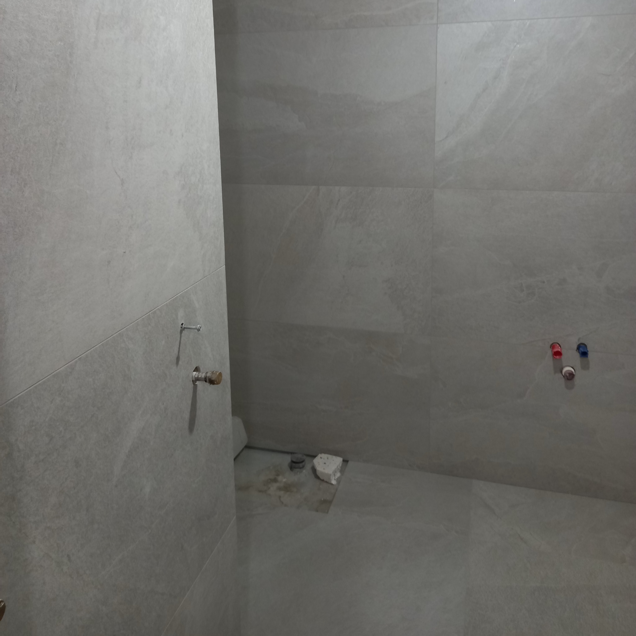 Wykończona łazienka z szarymi płytkami imitującymi kamień. Widoczne przyłącza wodne i fragment podłogi z pozostałościami po pracach glazurniczych.