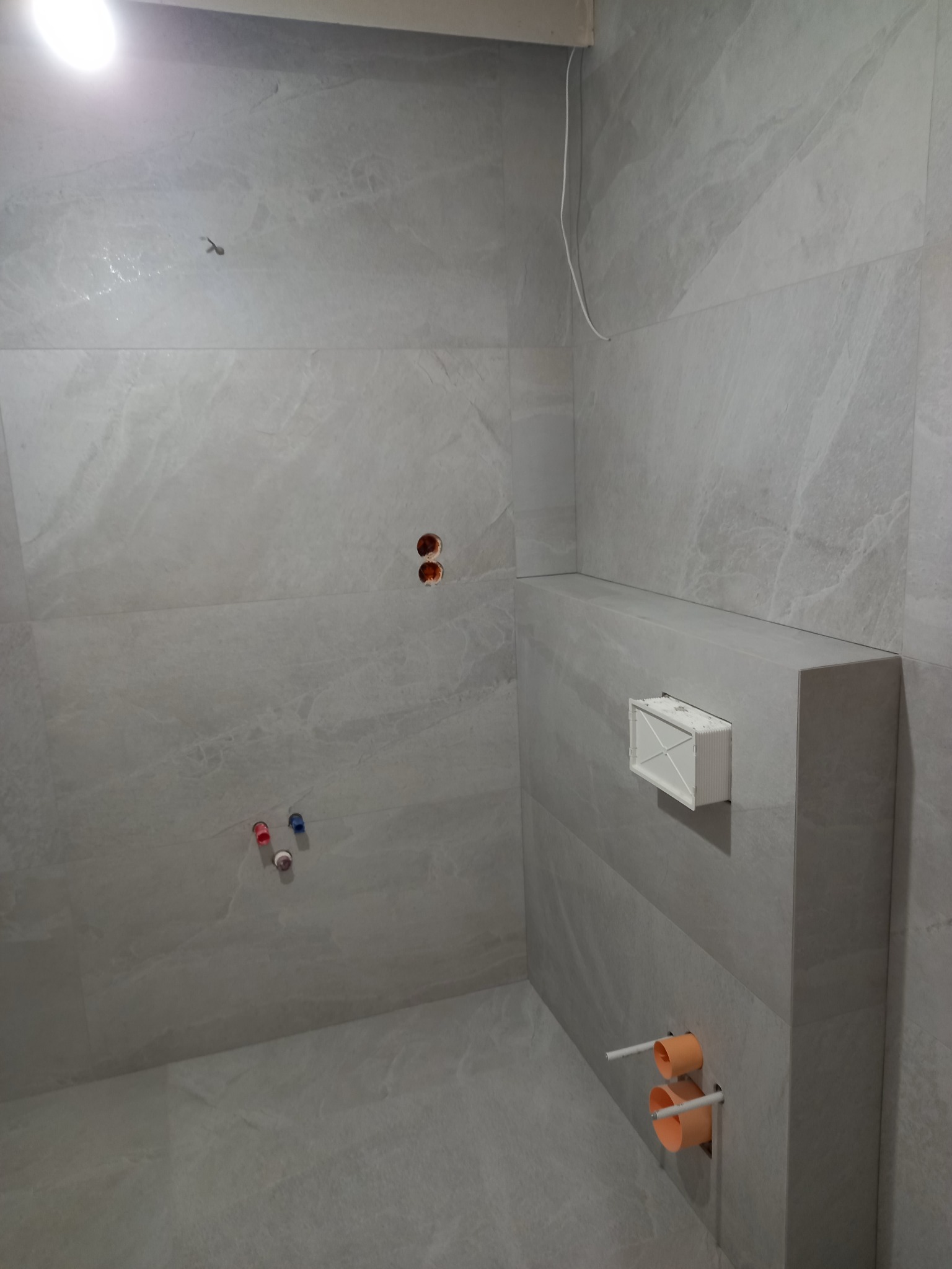 Wykończona łazienka z szarymi płytkami imitującymi kamień, widoczne otwory na armaturę i stelaż podtynkowy WC. Minimalistyczny styl.