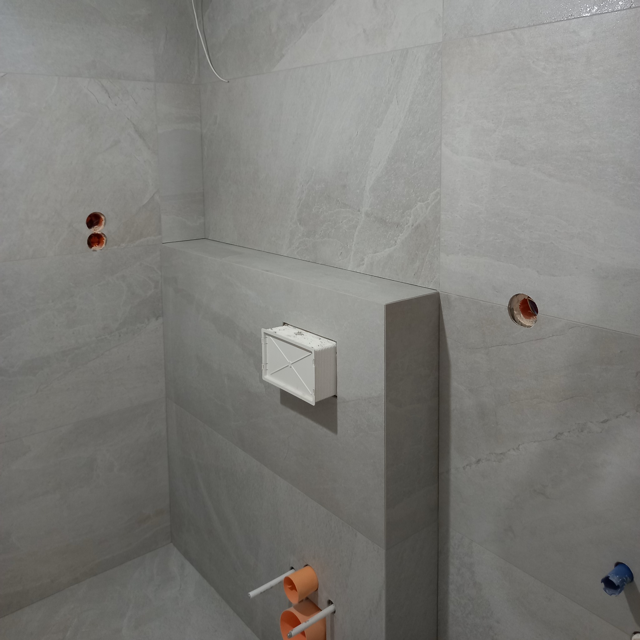 Wykończona łazienka z szarymi płytkami imitującymi marmur. Widoczne otwory na armaturę i zabudowany stelaż WC z białym rezerwuarem na papier toaletowy.