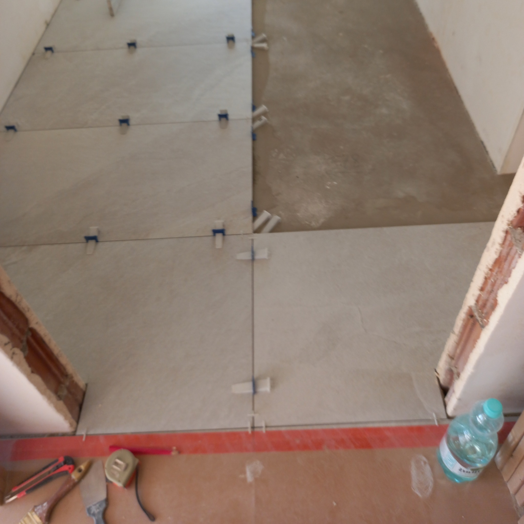 Układanie jasnych płytek podłogowych z użyciem poziomujących klipsów. Częściowo wykończona powierzchnia, widoczne narzędzia i butelka z wodą.