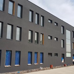 LUK-GOOD Patryk Lukoszek - Nowoczesny budynek z ciemnoszarą elewacją, okna zabezpieczone folią, widoczni pracownicy podczas prac wykończeniowych na zewnątrz.