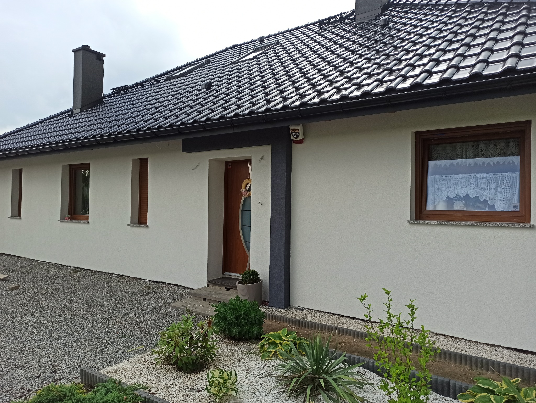 Nowoczesny dom jednorodzinny z białą elewacją o strukturze baranka, ciemną dachówką, drewnianymi oknami i drzwiami oraz alarmem na ścianie, otoczony ozdobnym ogrodem z kamykami i roślinami.