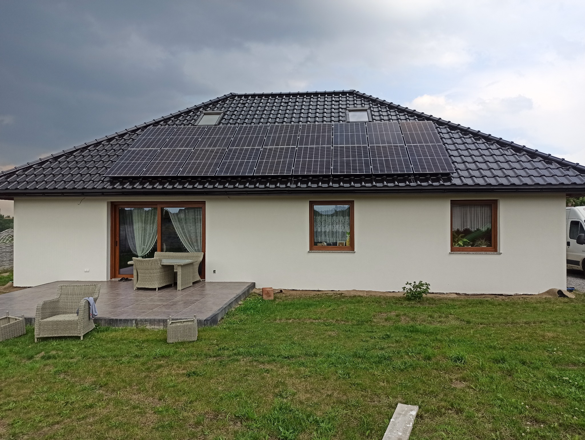 Jednorodzinny dom z jasną elewacją, ciemną dachówką, panelami słonecznymi, oknami w drewnianych ramach i tarasem z meblami ogrodowymi. W tle ciemne chmury.