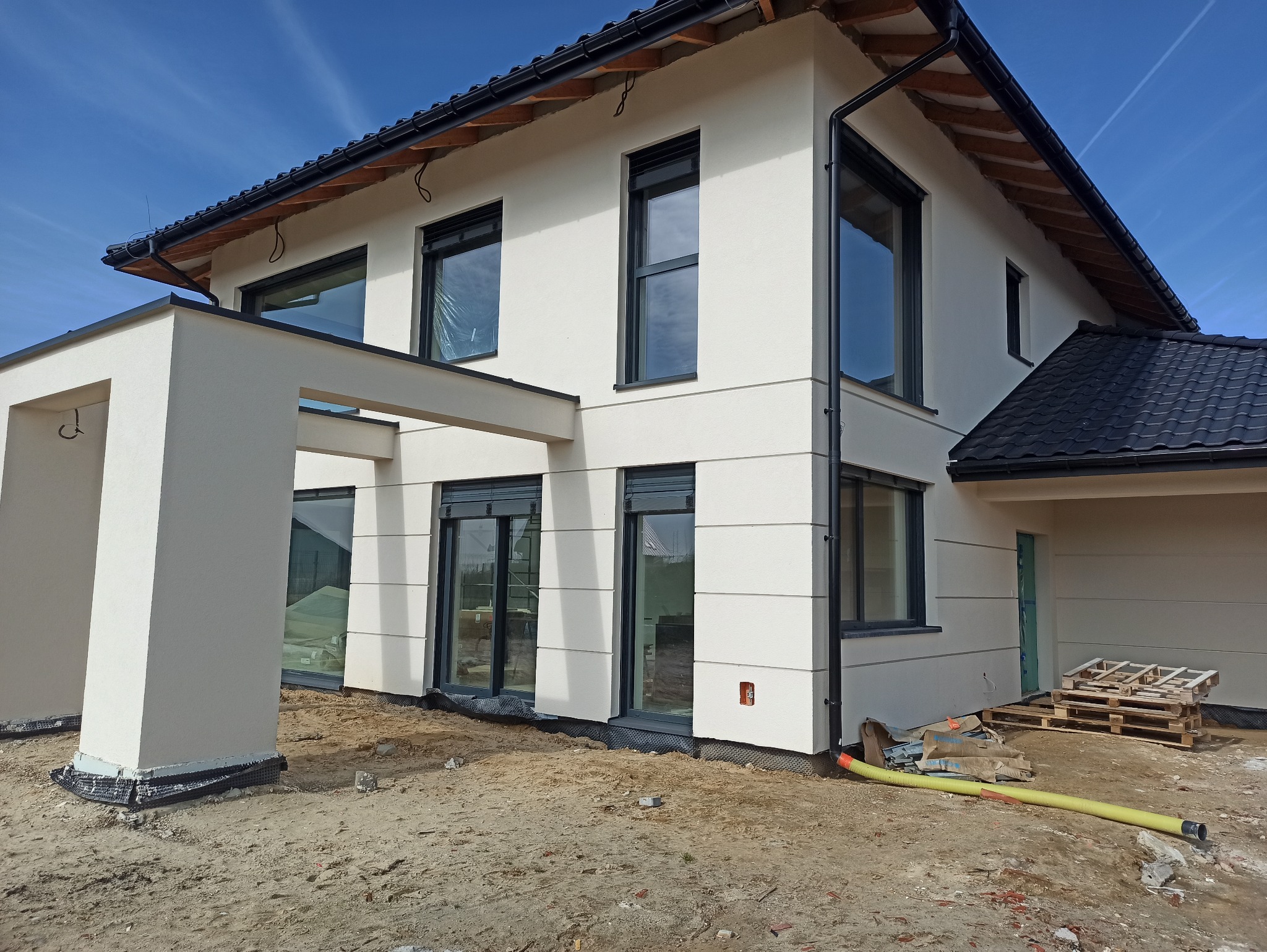 Nowoczesny dom jednorodzinny z jasną elewacją, czarną stolarką okienną i dachem z ciemnej dachówki, widoczne elementy wykończeniowe wokół budynku i żółty wąż na ziemi.