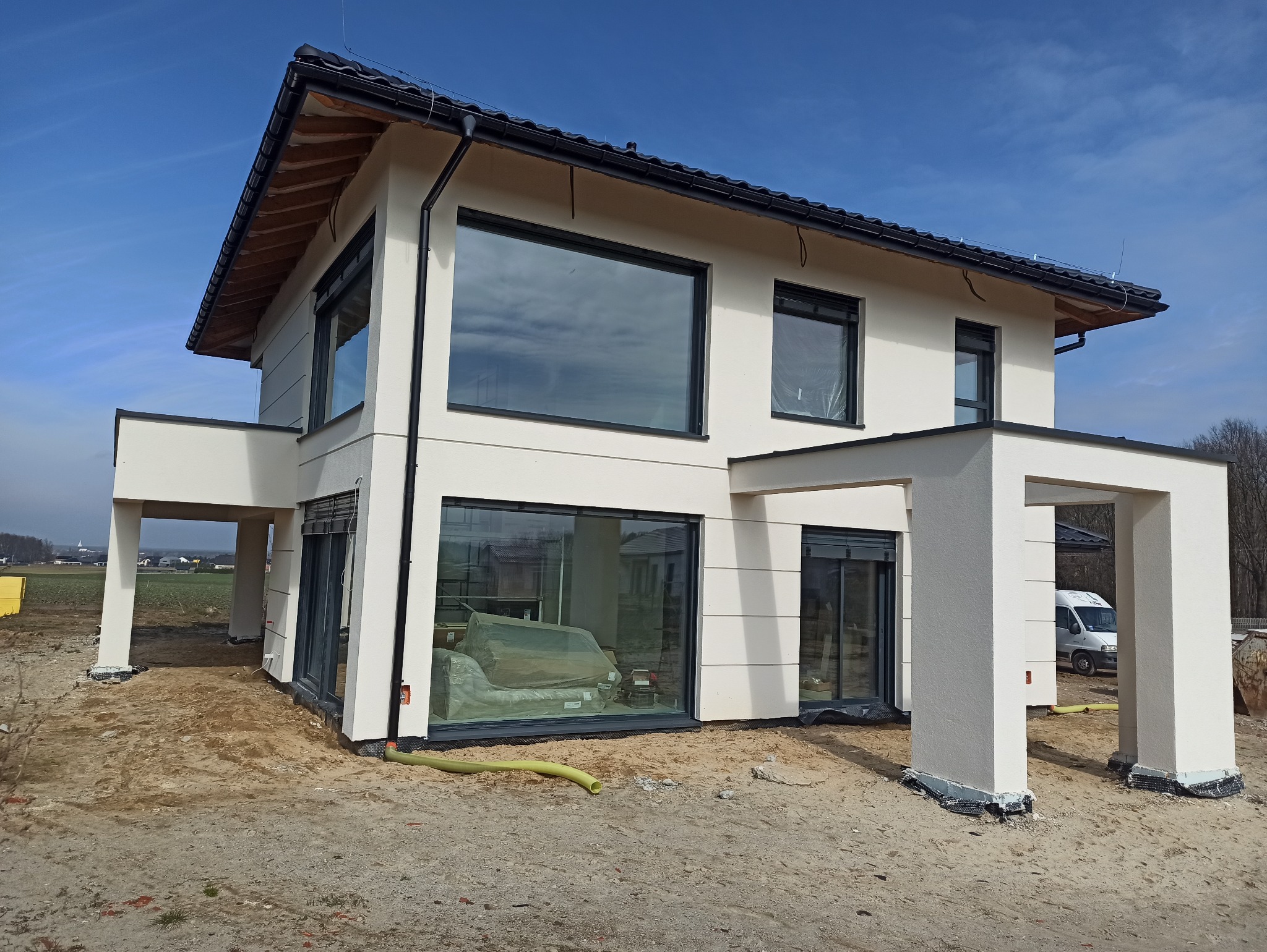Nowoczesny, dwukondygnacyjny dom z jasną elewacją i dużymi oknami, częściowo jeszcze zabezpieczonymi folią, widoczne elementy wykończeniowe przy fundamentach, w tle błękitne niebo i krajobraz.