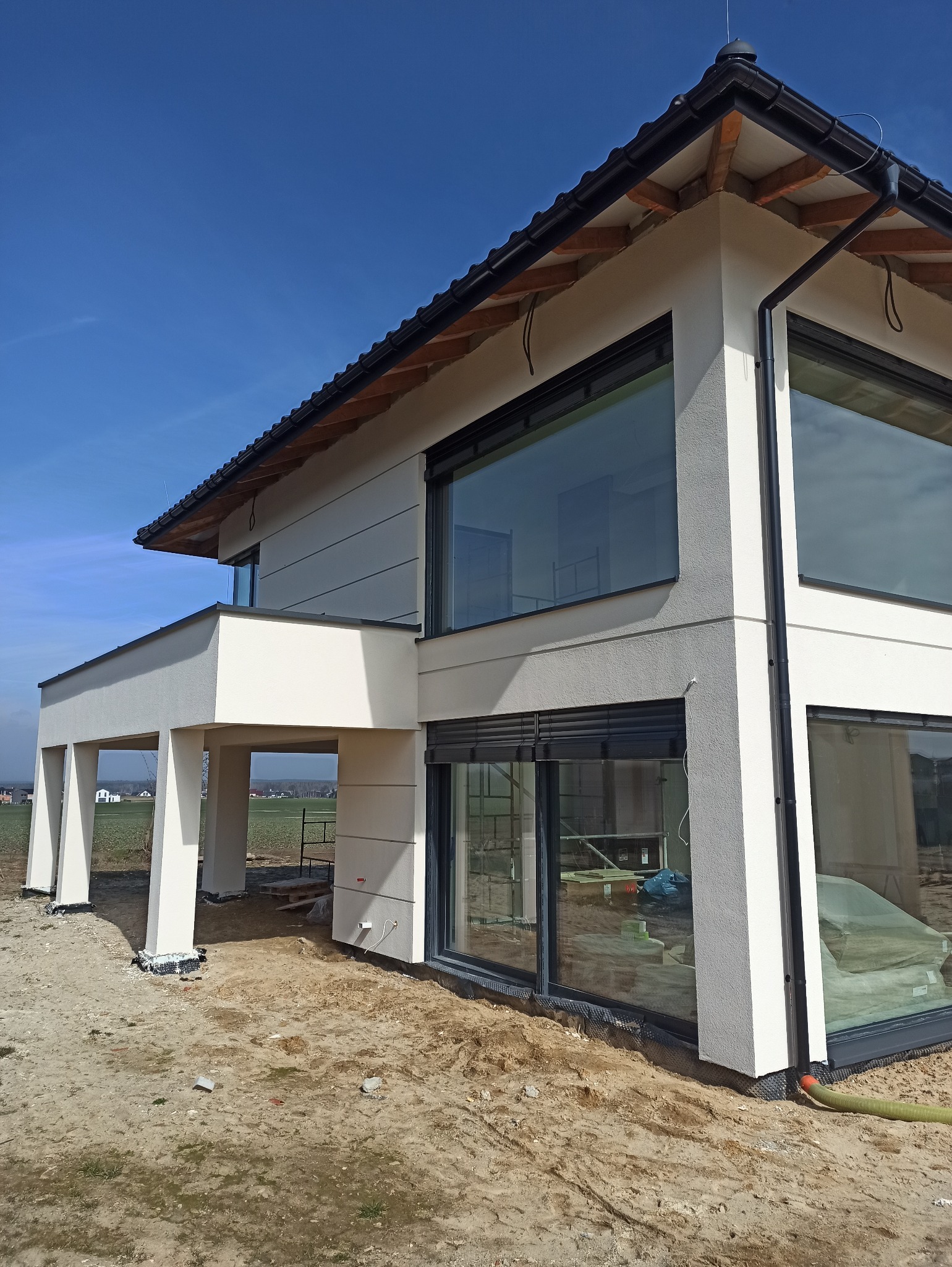 Nowoczesny dom jednorodzinny z dużą ilością przeszkleń i jasną elewacją, widoczny fragment ogrodu i niebo w tle, prace wykończeniowe w toku, teren wokół domu jeszcze niezagospodarowany.