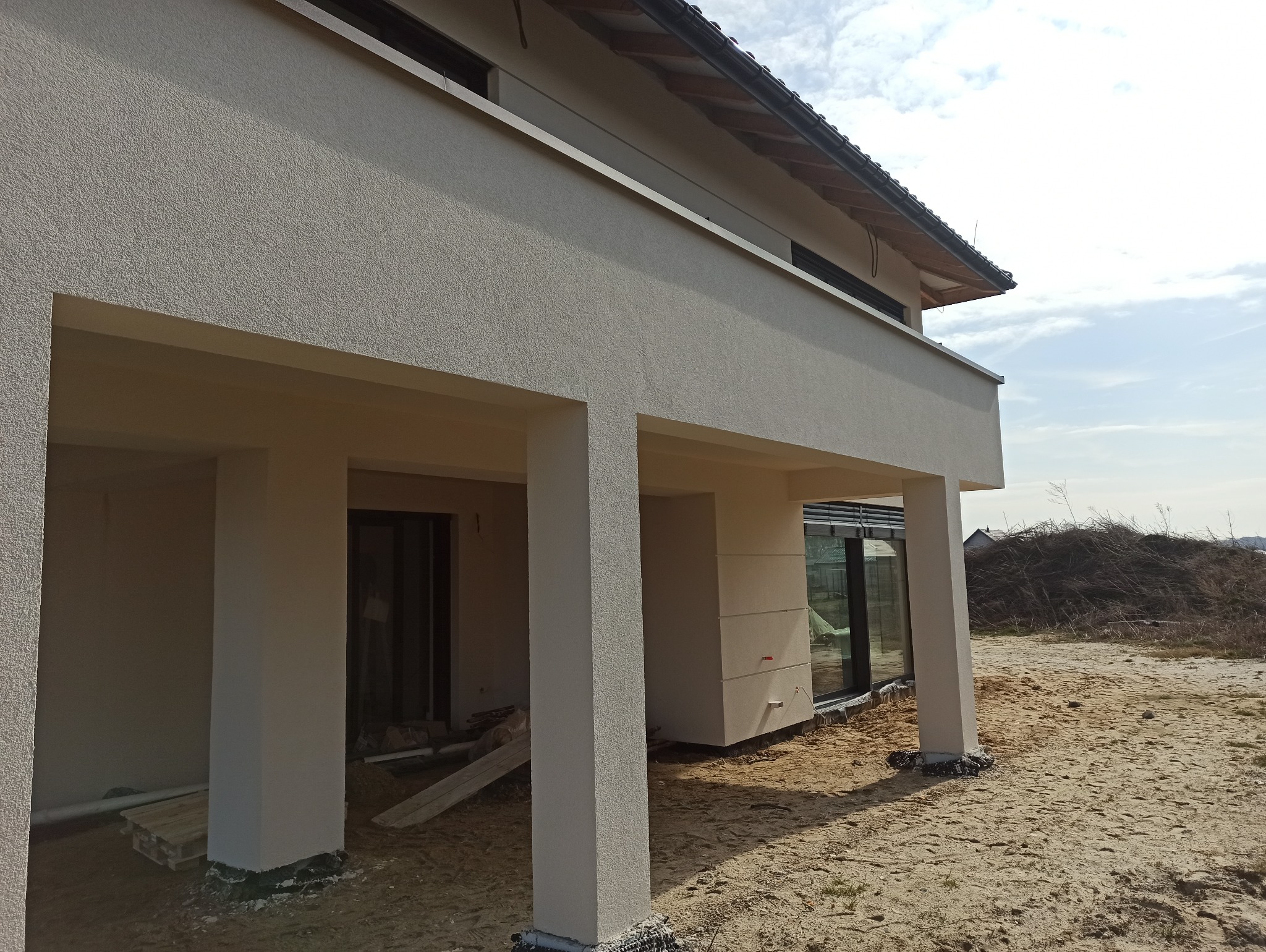 Nowoczesny dom w trakcie wykańczania elewacji, widoczne kolumny podtrzymujące zadaszenie, świeżo nałożony tynk i przygotowany teren wokół budynku z widocznym stosem gałęzi w tle.