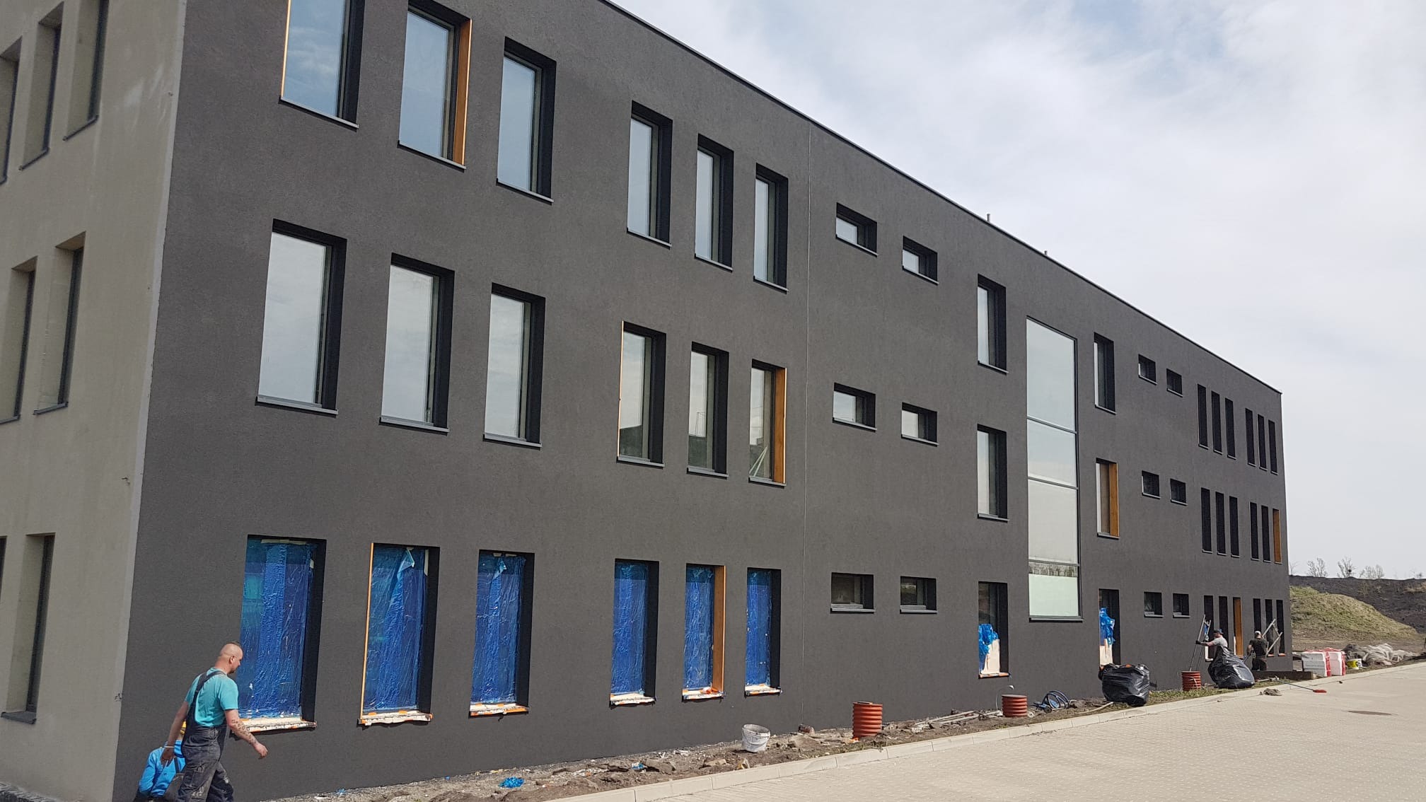 Nowoczesny budynek z ciemnoszarą elewacją, okna zabezpieczone folią, widoczni pracownicy podczas prac wykończeniowych na zewnątrz.