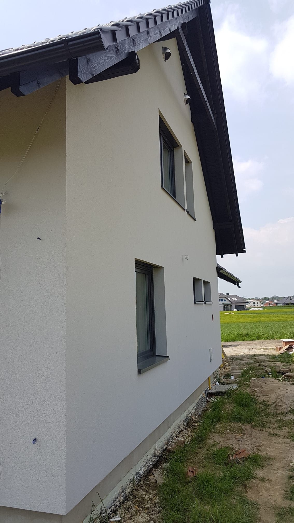 Boczny widok na nowo wykończony dom z jasną elewacją o delikatnej fakturze, ciemnoszarą stolarką okienną i czarnym dachem z drewnianymi elementami dekoracyjnymi; widoczny fragment trawnika...