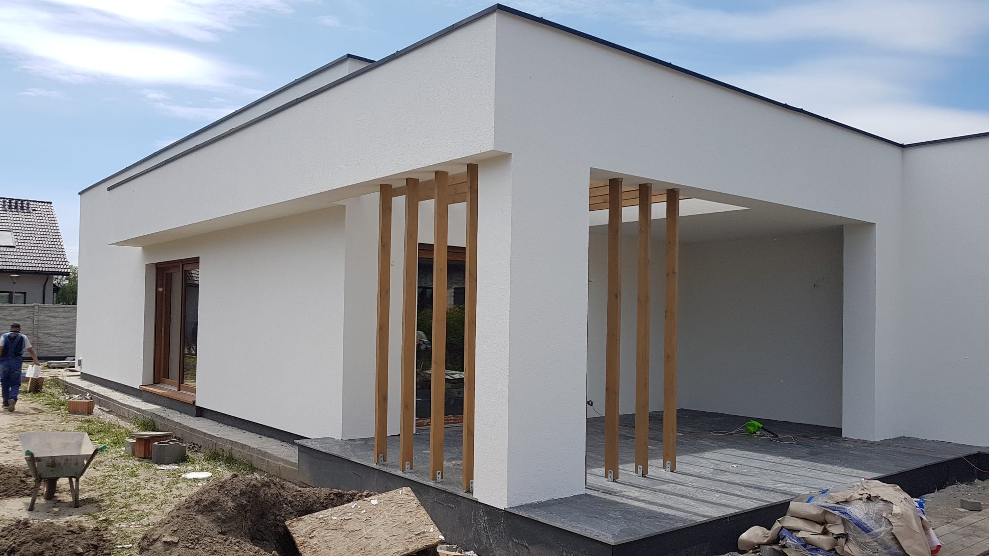 Nowoczesny dom jednorodzinny z białą elewacją i drewnianymi elementami dekoracyjnymi, widoczny w trakcie prac wykończeniowych, z workami zaprawy i taczką na pierwszym planie.