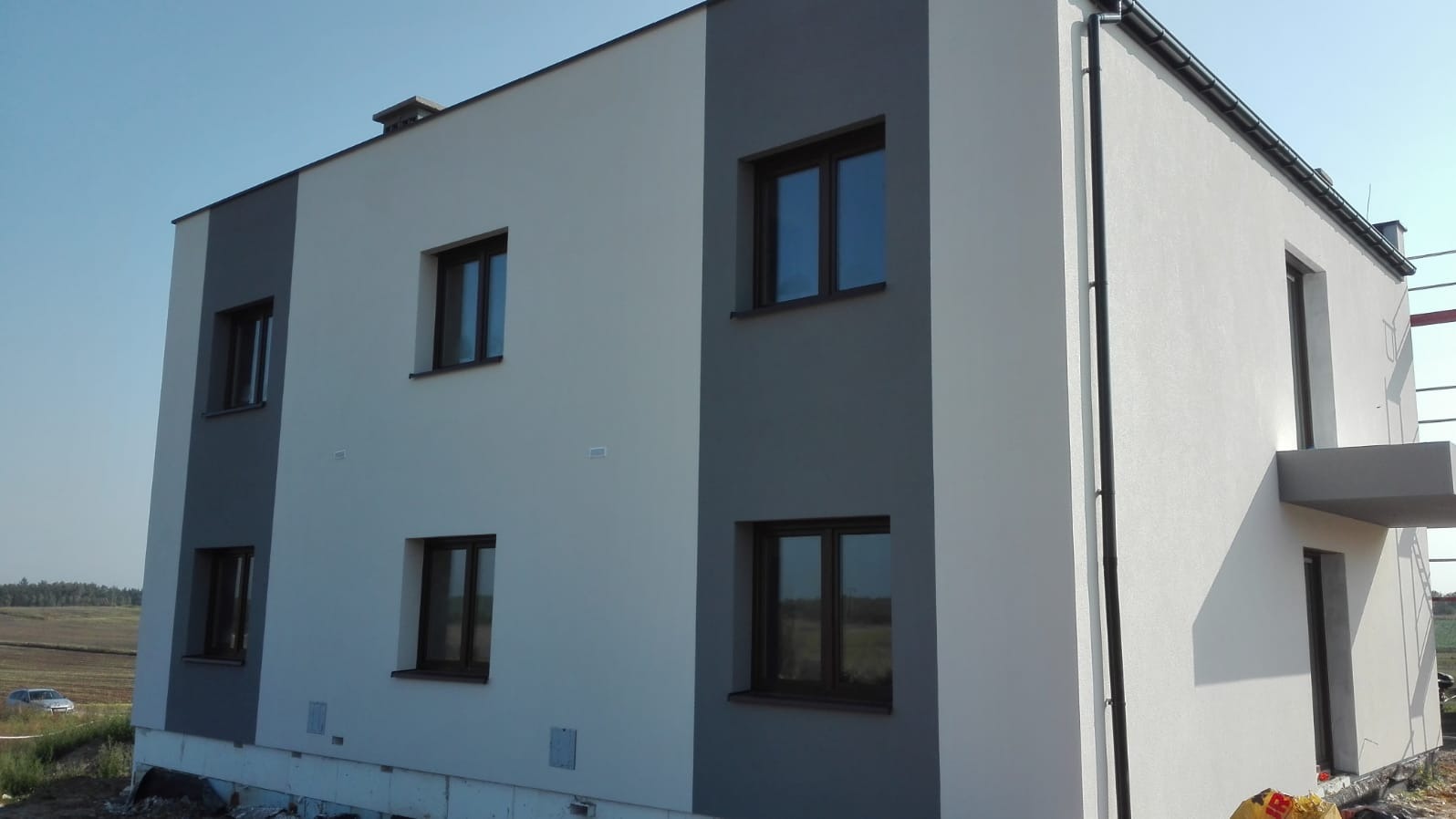 Dwukondygnacyjny budynek z elewacją w odcieniach szarości i bieli, z ciemnymi oknami, widok z dołu ukazujący częściowo wykończone otoczenie.