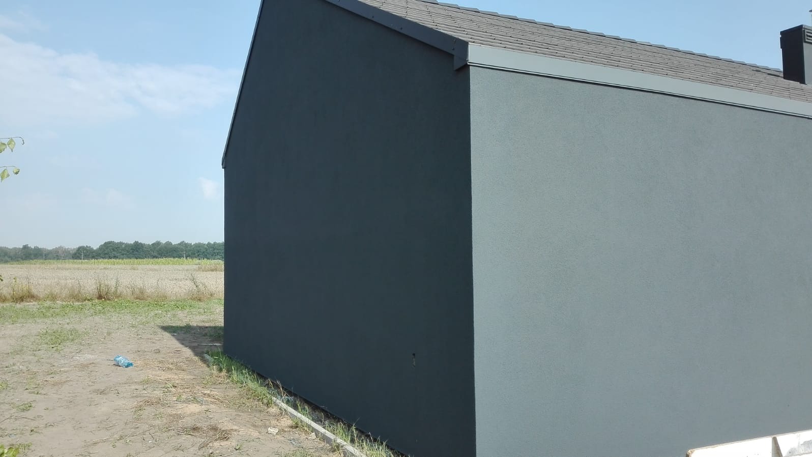 Nowoczesny budynek z szarą elewacją i ciemnoszarą dachówką na tle pola i błękitnego nieba, widoczne ślady lekkiego zabrudzenia na elewacji przy ziemi oraz pozostawiona butelka plastikowa obok budynku.