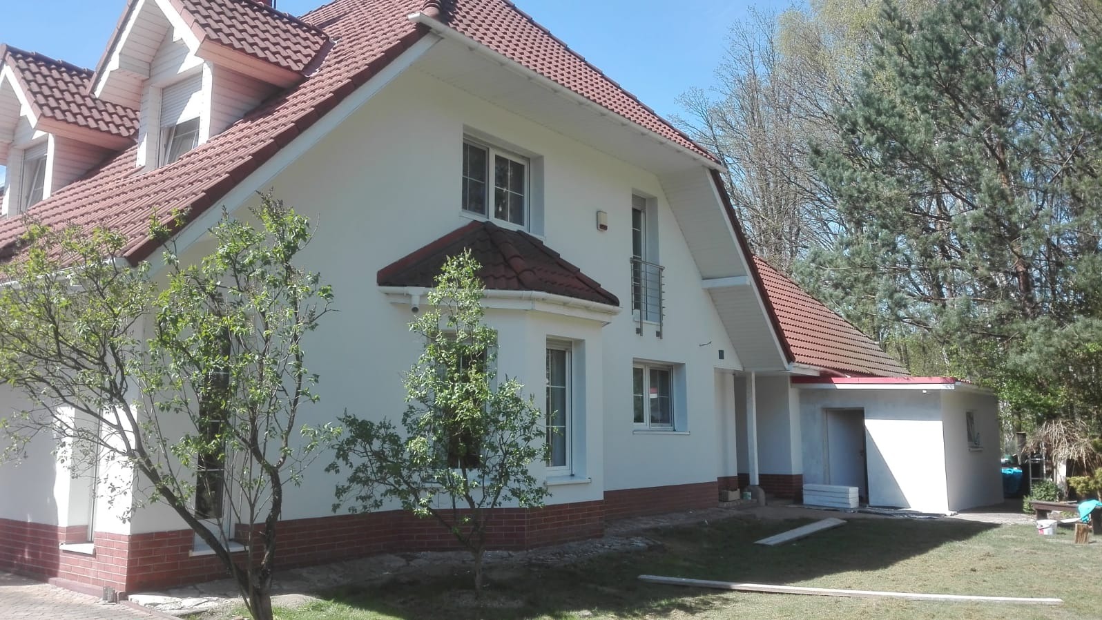 Wykończona elewacja domu jednorodzinnego z czerwonym dachem, widoczne białe ściany, okna i drzewa w ogrodzie, garaż dobudowany do domu.