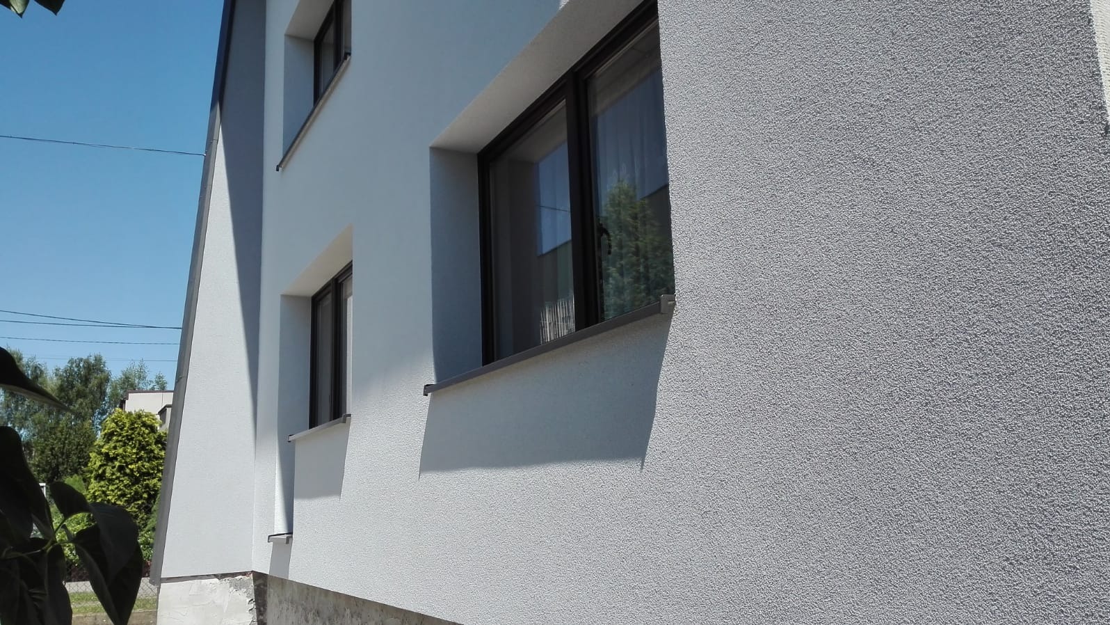 Fragment ściany domu jednorodzinnego z oknami w ciemnych ramach, po wykonaniu nowej, szarej elewacji z wyraźną fakturą baranka, widok z dołu.