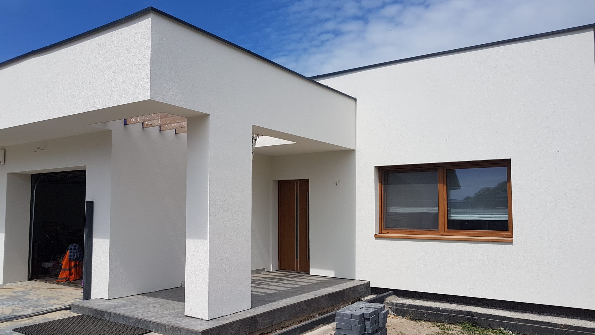 Nowoczesny dom jednorodzinny z białą elewacją, drewnianą stolarką okienną i minimalistycznym wejściem z kolumnami, widoczne kostki brukowe przed wejściem.