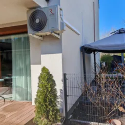 Zamontowana jednostka zewnętrzna klimatyzacji Rotenso Versu na elewacji domu, z widocznym odprowadzeniem skroplin i małym drzewkiem iglastym u dołu ściany.