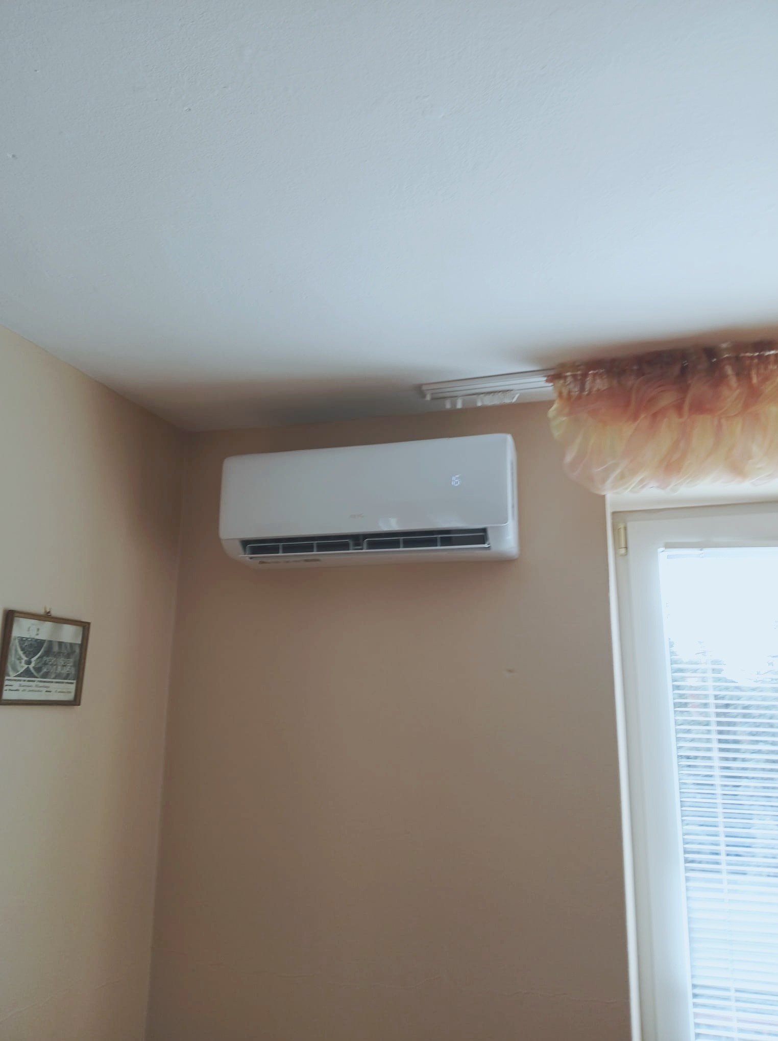 Nowoczesny, biały klimatyzator ścienny zamontowany w pokoju z beżowymi ścianami, obok okna z żaluzjami i dekoracyjną zasłoną.