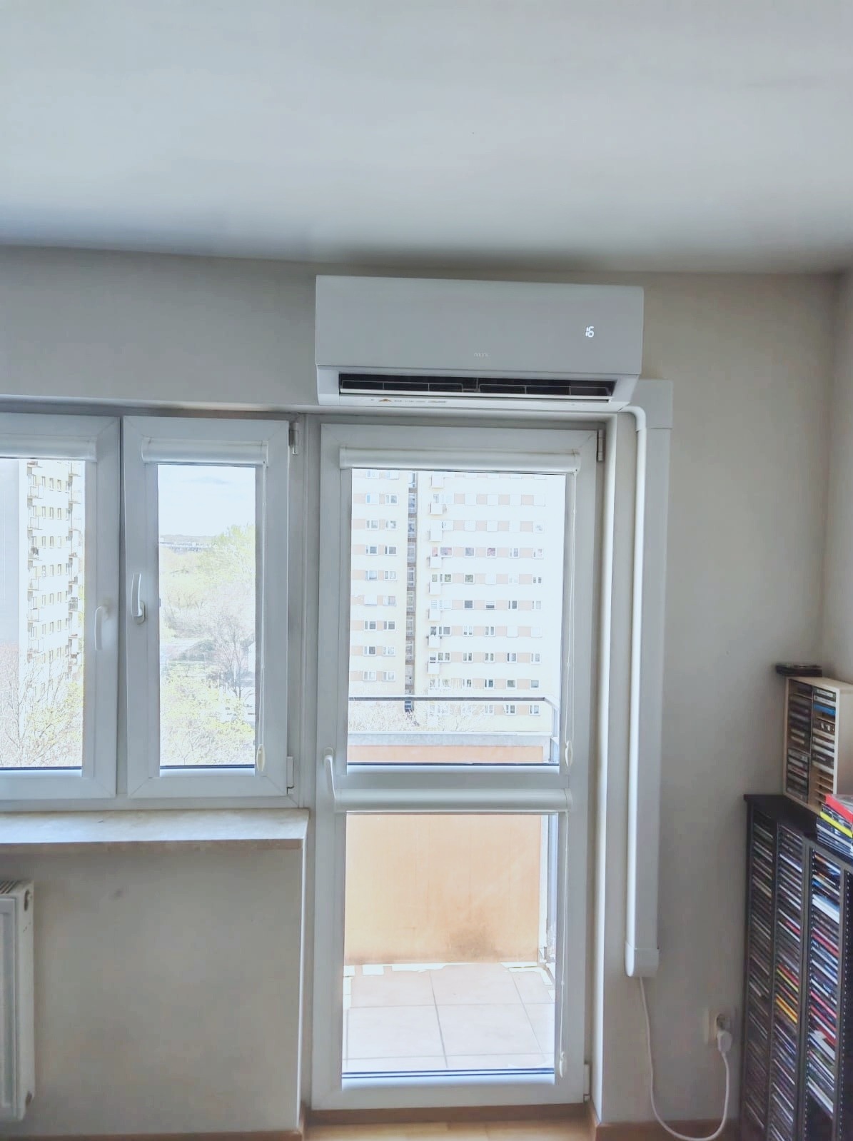 Biały klimatyzator na ścianie nad oknem balkonowym w mieszkaniu. Widok na blok z balkonu. Na klimatyzatorze wyświetla się temperatura 16 stopni.