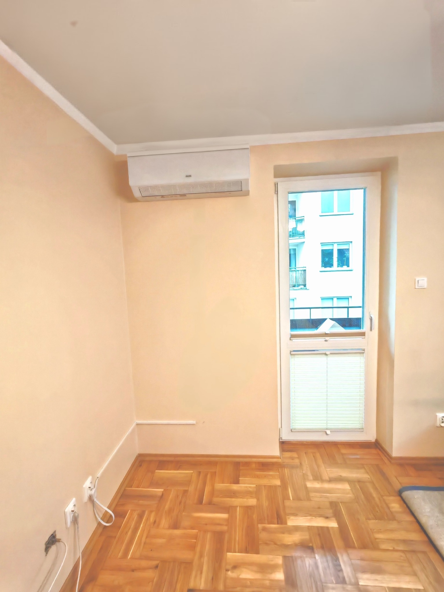 Wnętrze z zamontowaną klimatyzacją AUX nad oknem balkonowym w Karczewie. Widoczna drewniana podłoga i jasne ściany.