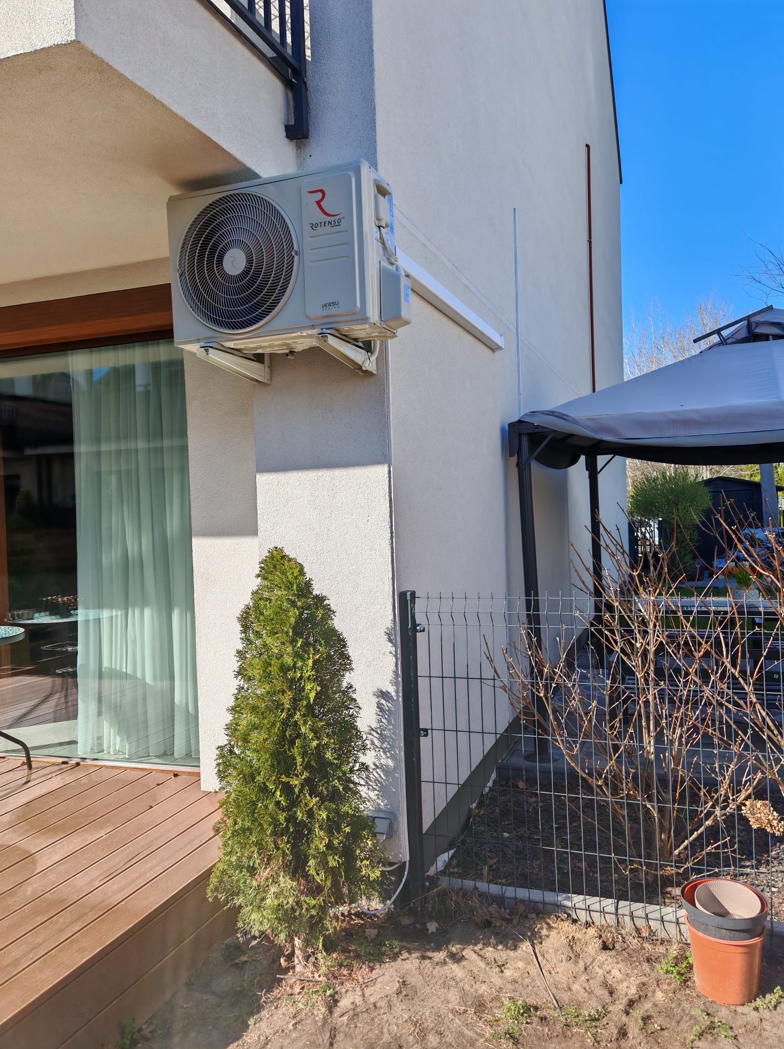 Zamontowana jednostka zewnętrzna klimatyzacji Rotenso Versu na elewacji domu, z widocznym odprowadzeniem skroplin i małym drzewkiem iglastym u dołu ściany.