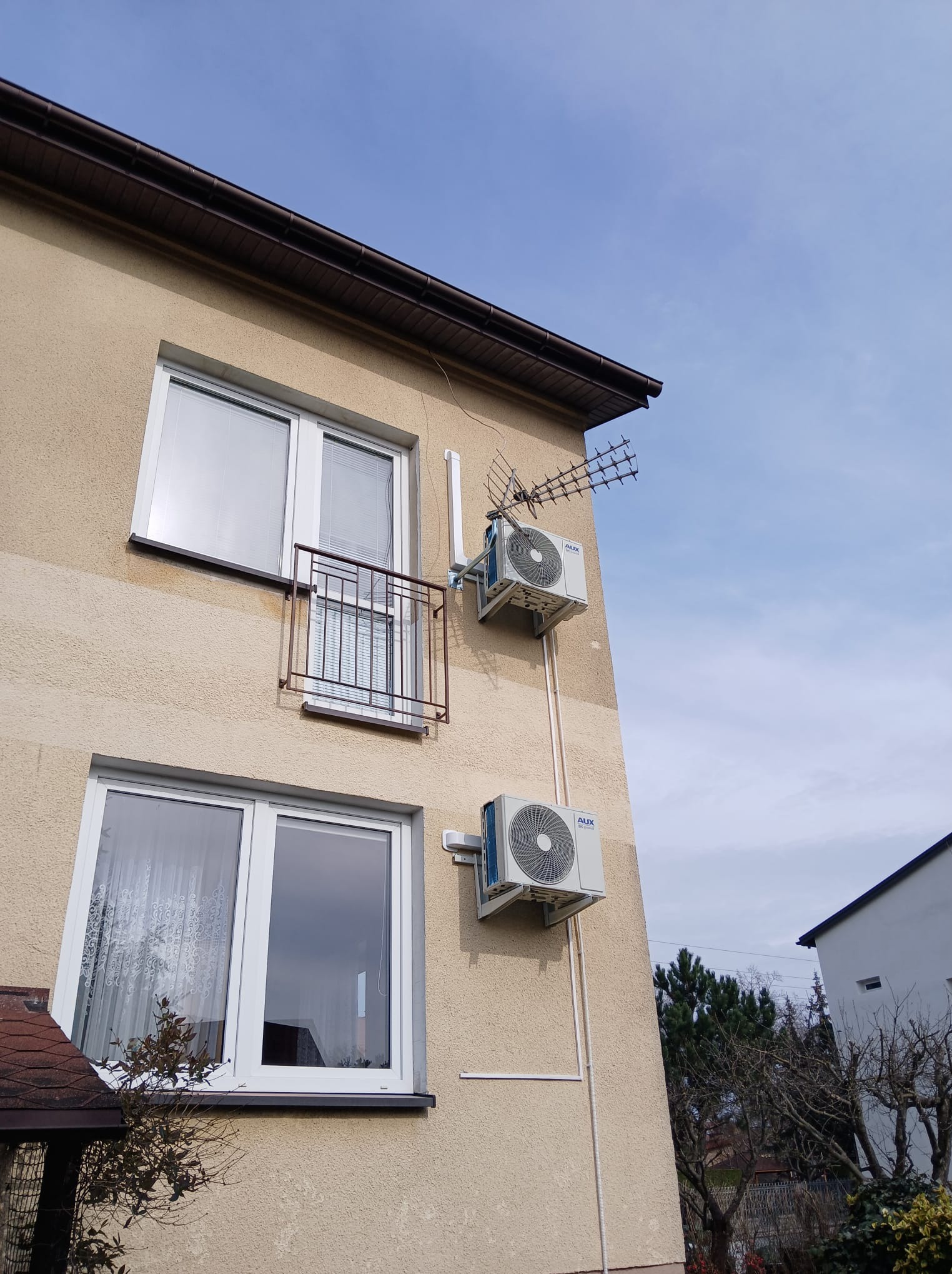 Elewacja budynku z dwoma zamontowanymi klimatyzatorami marki AUX, jeden z anteną TV. Widoczne okna i balkon. Jasne niebo w tle.