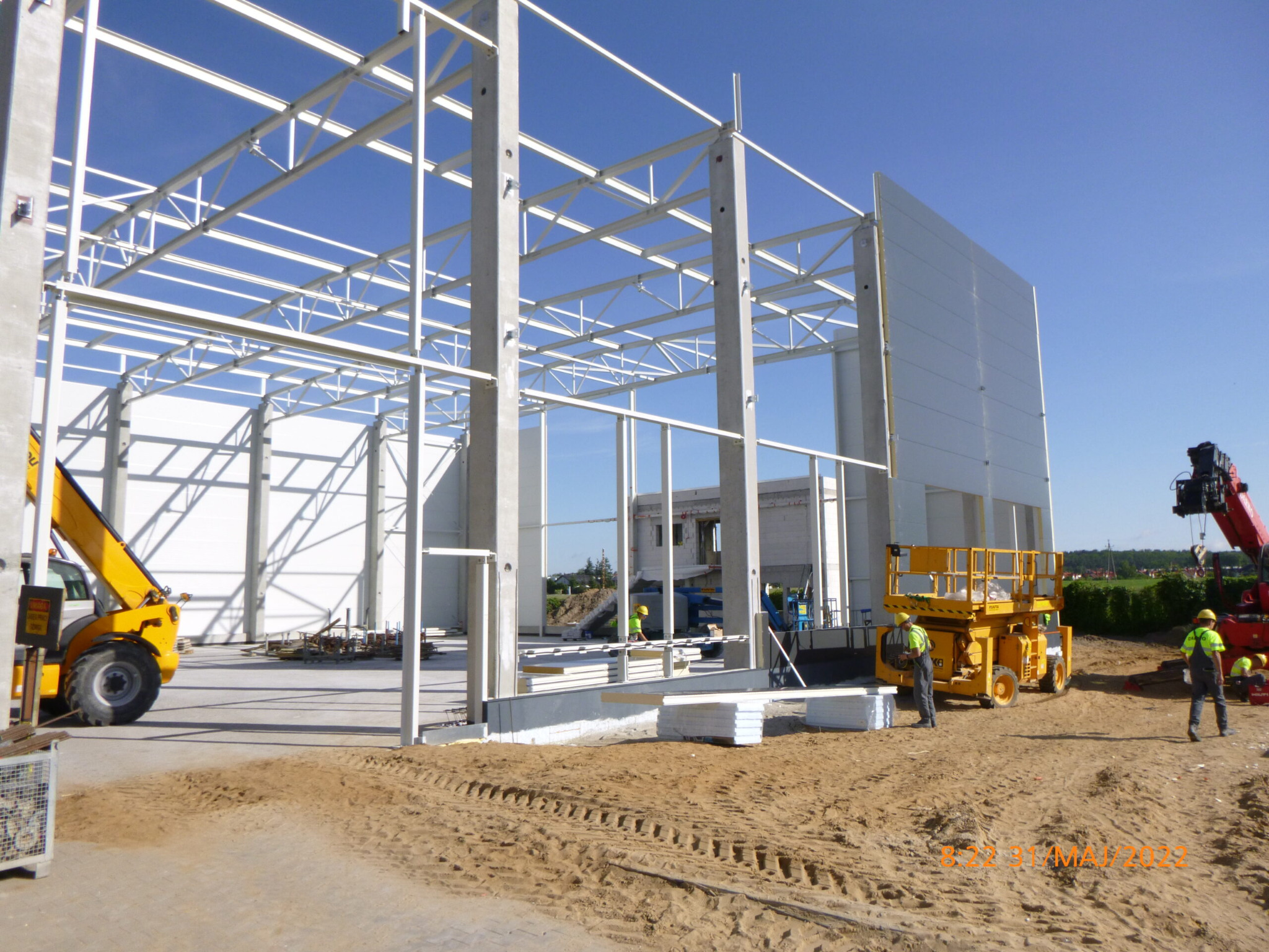 Montaż konstrukcji stalowej hali z wykorzystaniem podnośników i dźwigu, widoczne betonowe słupy i białe panele ścienne.