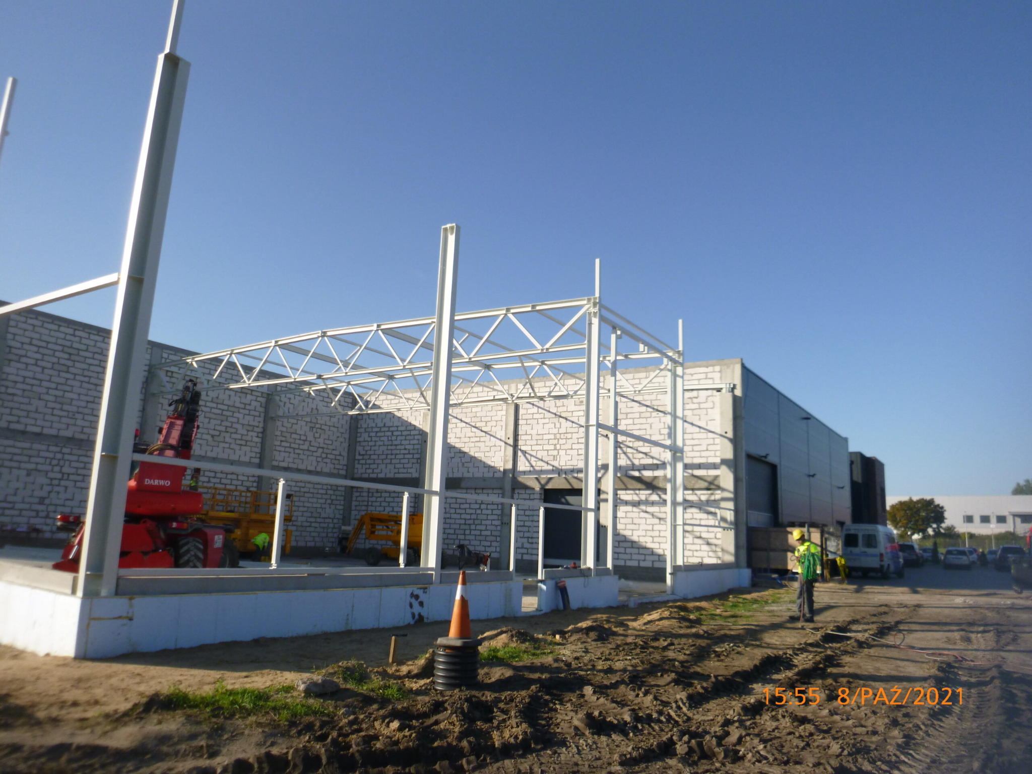 Wznoszenie konstrukcji stalowej hali z elementami murowanymi, widoczny czerwony podnośnik DARWO i pracownik w odblaskowej kamizelce na placu budowy.