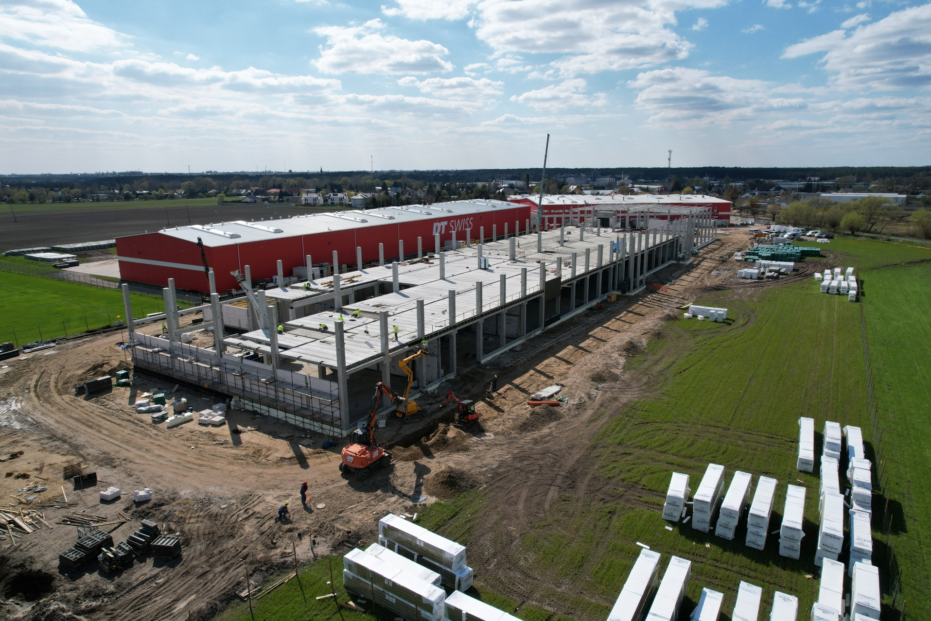 Wysoka perspektywa na budowę hali z elementami żelbetowymi, widoczny szkielet konstrukcji, pracujący robotnicy i maszyny budowlane, obok gotowy budynek z logo DT Swiss.