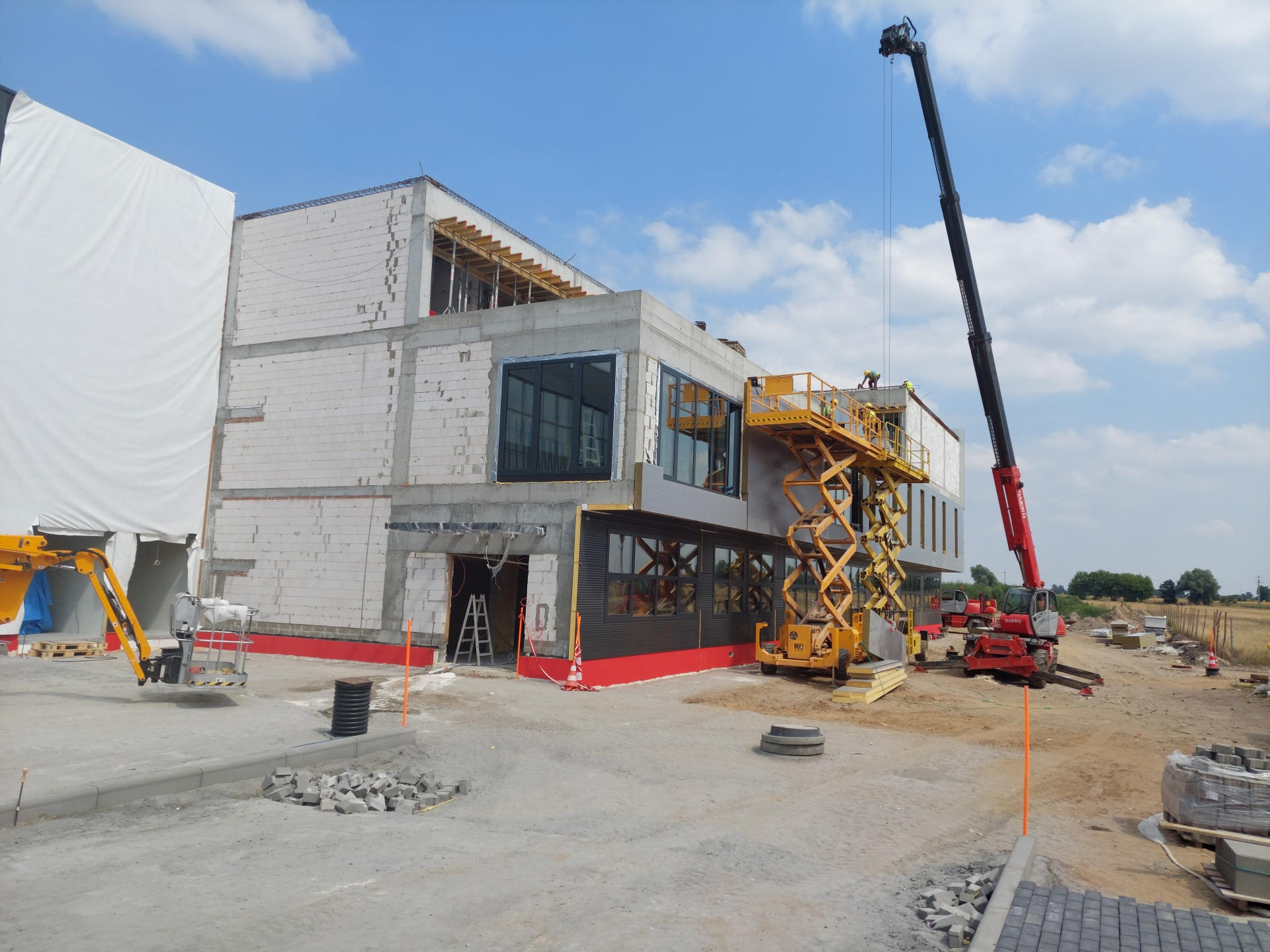 Wznoszenie dwukondygnacyjnego budynku z betonu komórkowego z elementami konstrukcji stalowej, widoczny żuraw teleskopowy oraz podnośnik nożycowy, teren budowy w trakcie prac wykończeniowych.