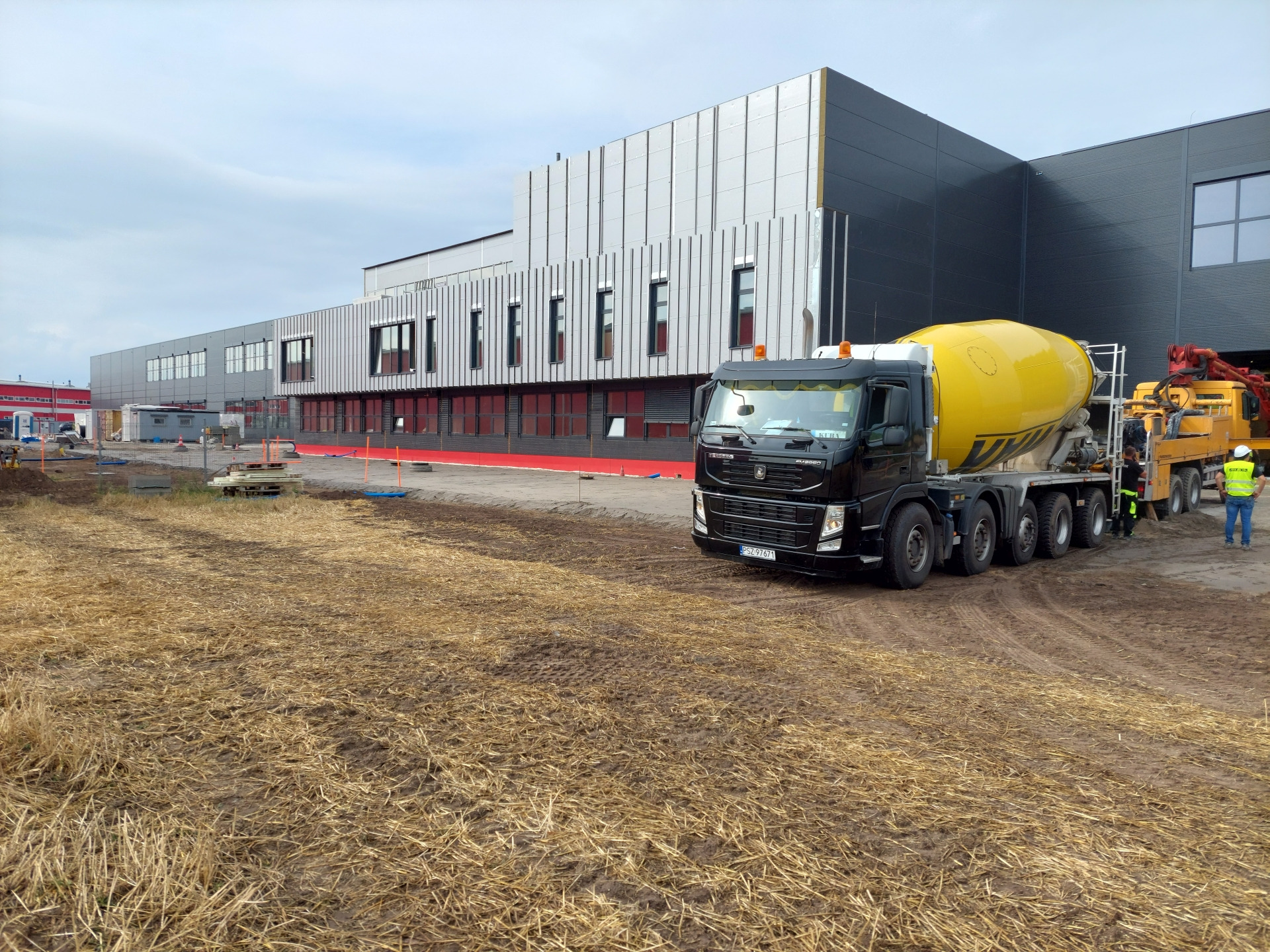 Ciężarówka z betoniarką na placu budowy nowoczesnego budynku przemysłowego z szarymi panelami elewacyjnymi i czerwonymi akcentami, widoczne przygotowane podłoże.