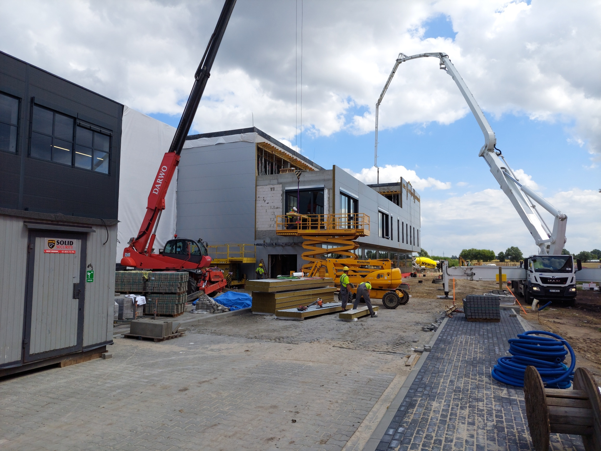 Montaż paneli ściennych na nowo budowanym obiekcie z użyciem podnośnika nożycowego i żurawia, widoczna betonomieszarka w tle.