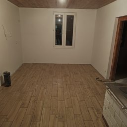 Usługi remontowo budowlane - Ułożone panele podłogowe imitujące drewno w pustym pokoju z drewnianym sufitem, widoczne gniazdka elektryczne i przewody, fragment pieca kaflowego w rogu.