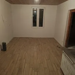 Ułożone panele podłogowe imitujące drewno w pustym pokoju z drewnianym sufitem, widoczne gniazdka elektryczne i przewody, fragment pieca kaflowego w rogu.