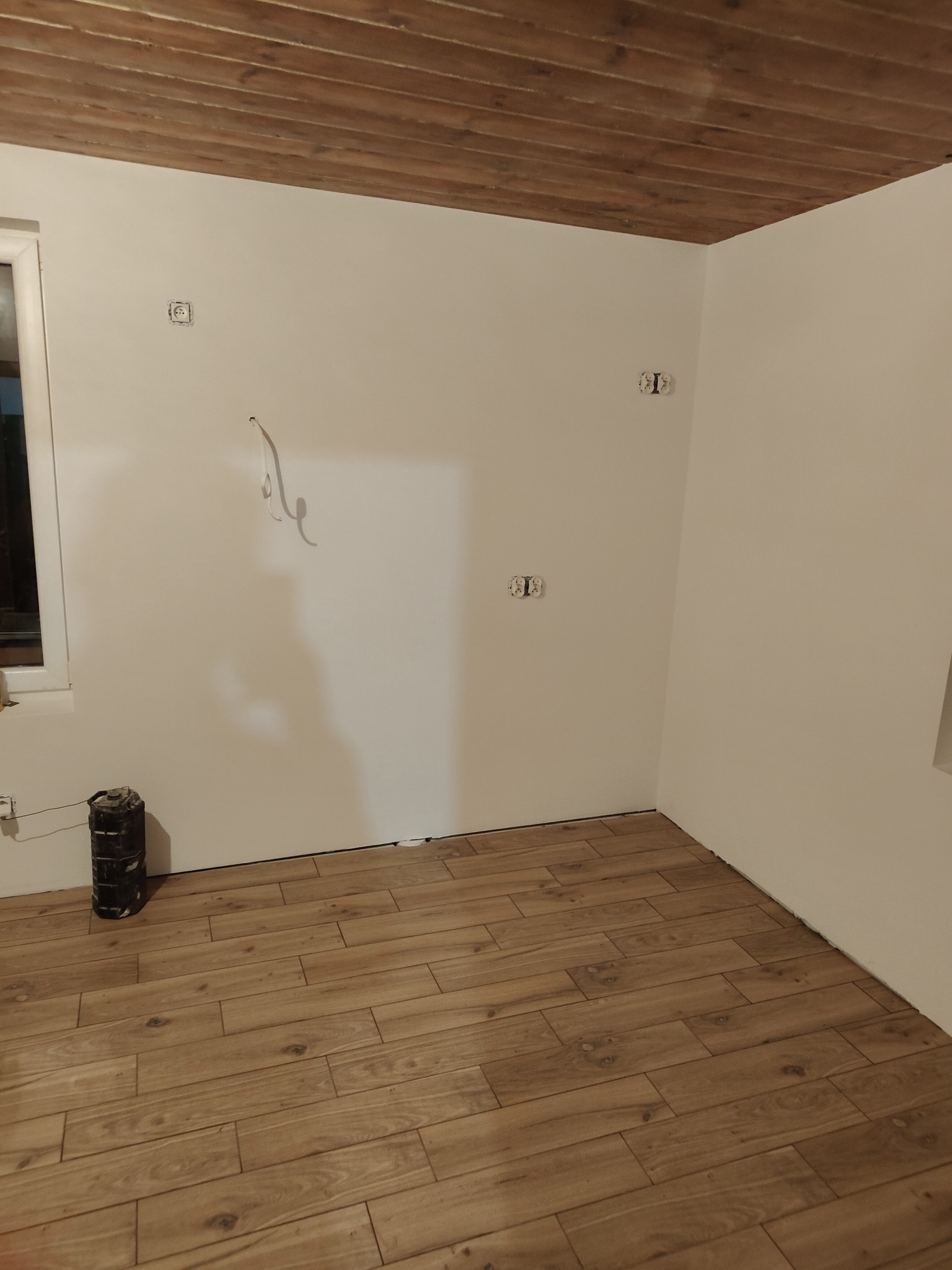Wnętrze pokoju z drewnianą podłogą i sufitem, świeżo pomalowane ściany z wystającymi przewodami elektrycznymi i puszkami gniazdek, widoczna puszka instalacyjna przy ścianie, okno po lewej stronie.