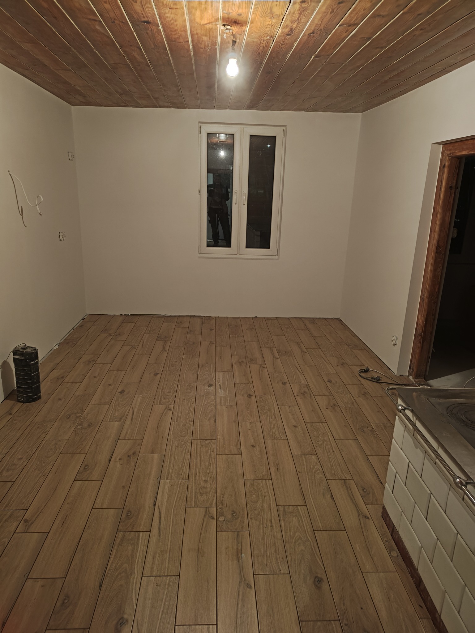 Ułożone panele podłogowe imitujące drewno w pustym pokoju z drewnianym sufitem, widoczne gniazdka elektryczne i przewody, fragment pieca kaflowego w rogu.
