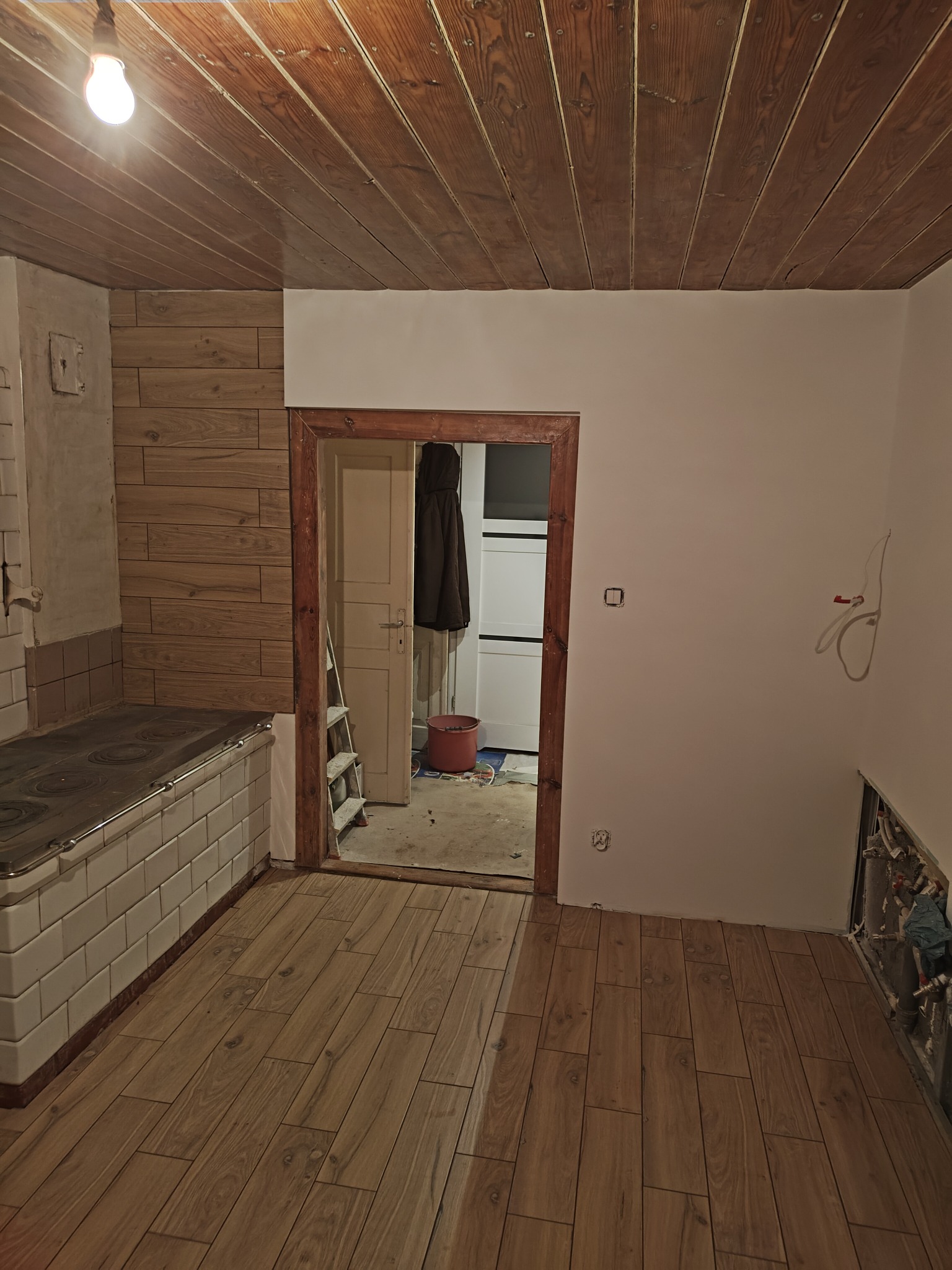 Wnętrze w trakcie remontu: drewniana boazeria na suficie i ścianie, nowa podłoga z paneli imitujących drewno, stara kuchnia kaflowa, instalacje elektryczne i hydrauliczne częściowo odsłonięte...