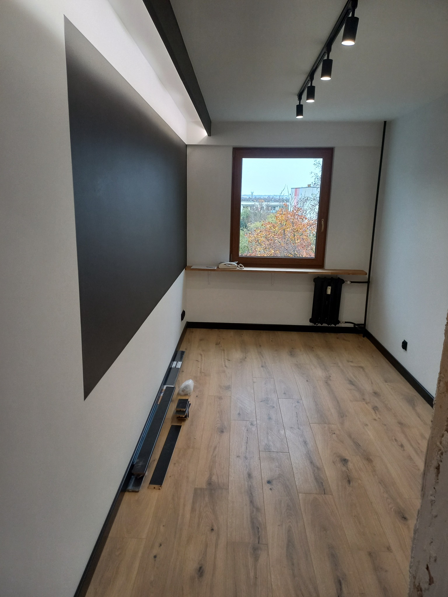 Pokój po remoncie z drewnianą podłogą, czarno-białą ścianą z oświetleniem LED, widokiem z okna na drzewa i grzejnikiem pod parapetem. Na podłodze leżą listwy przypodłogowe.