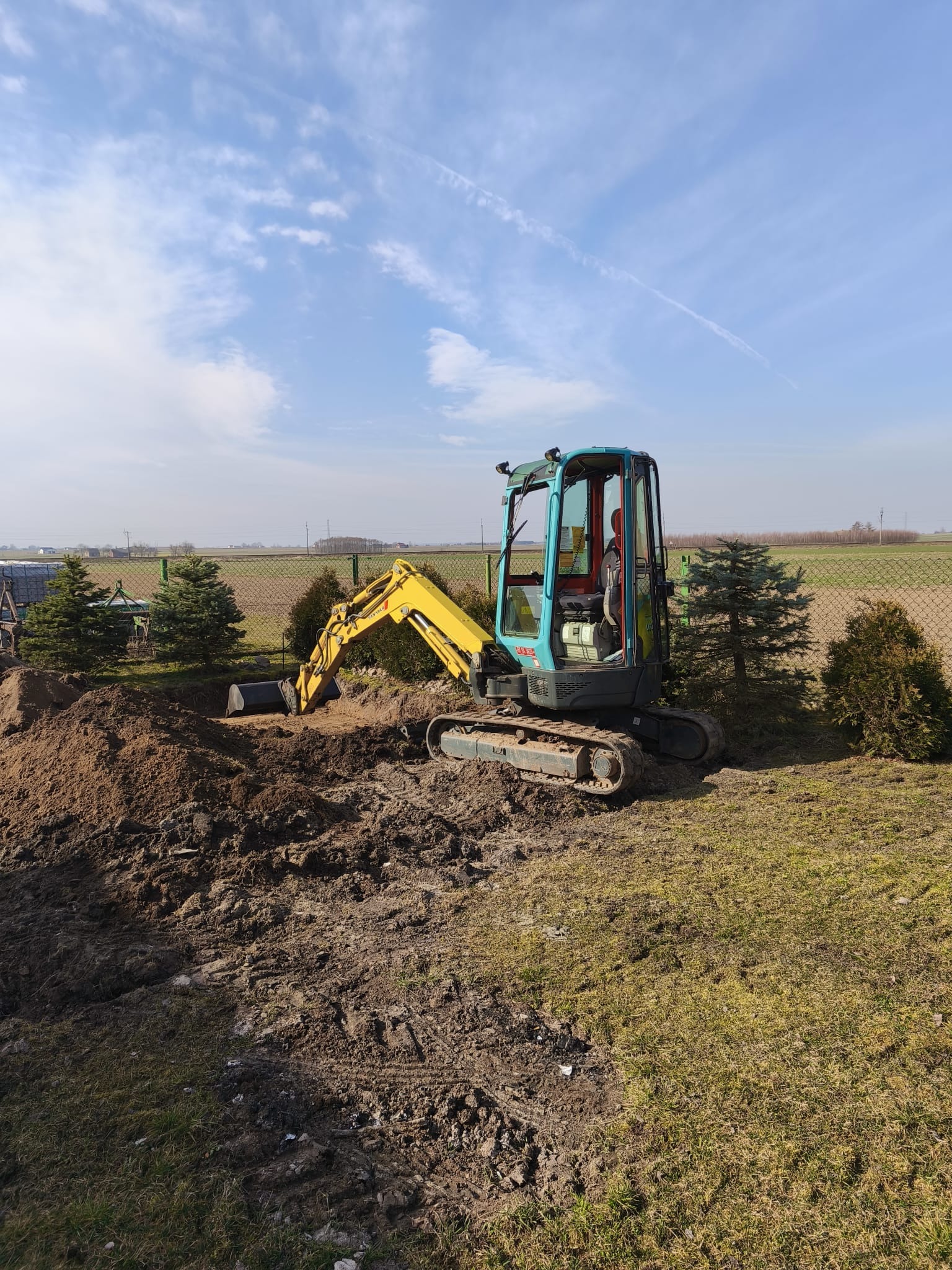 Mała koparka Yanmar, z żółtym ramieniem i turkusową kabiną, wykonuje wykop w ziemi. W tle widoczne zielone drzewka, siatka ogrodzeniowa i pole uprawne.